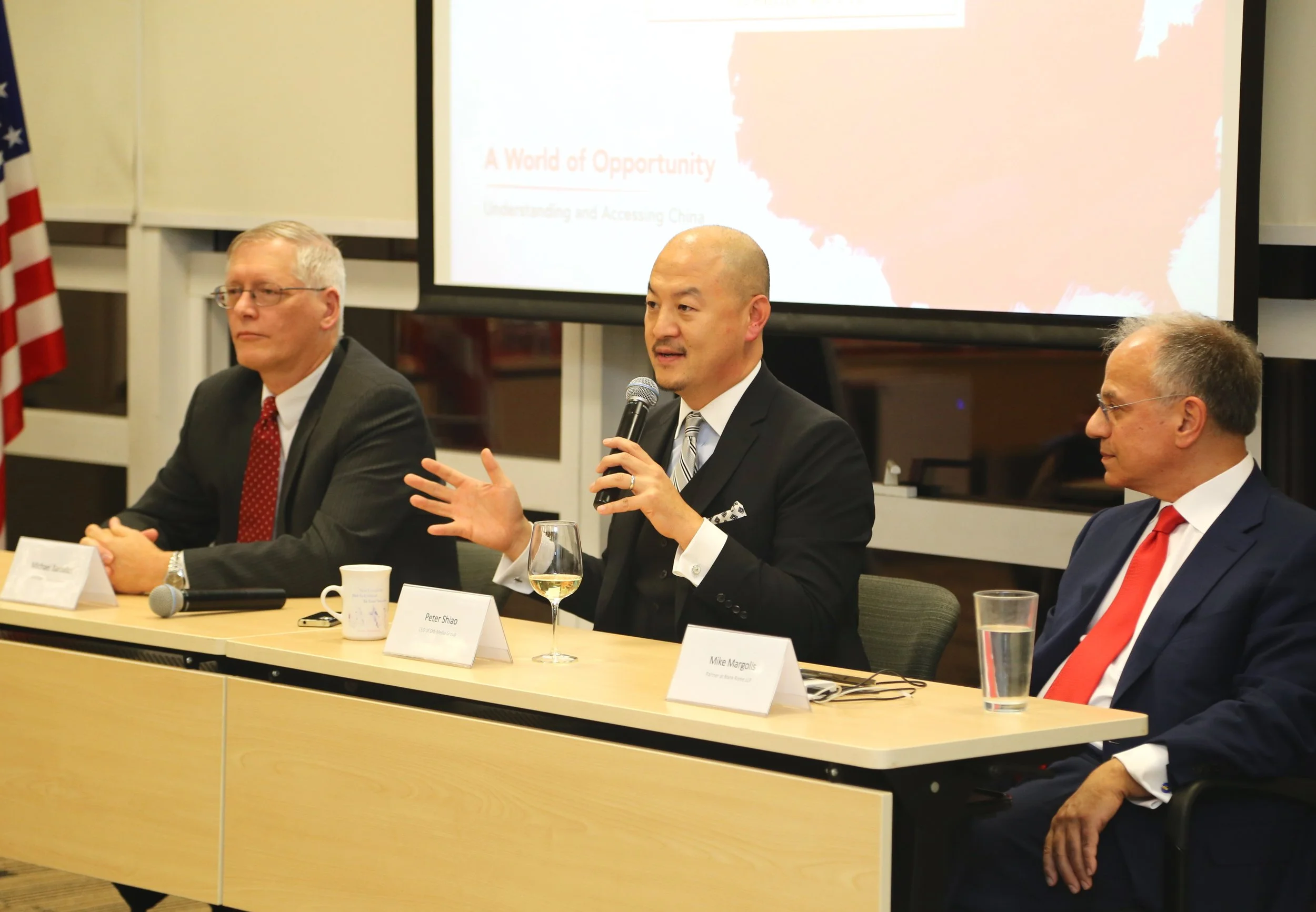 A panel of three men sitting at a conference table, with one man speaking into a microphone. The man in the center is holding the microphone, gesturing with his hand, and has a glass of white wine in front of him. The other two men are listening, with water and coffee cups on the table. A presentation screen behind them displays the title 'A World of Opportunity'.