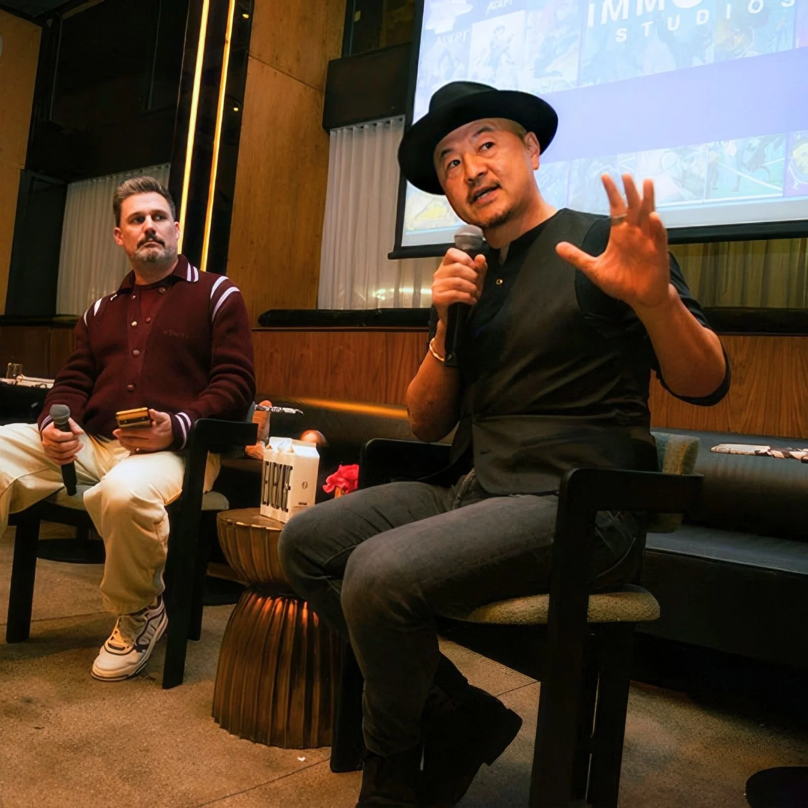 Two men seated on chairs participating in a discussion or presentation at an indoor event. The man on the right, wearing a black hat and holding a microphone, is speaking. The man on the left is holding a microphone and looking attentively.