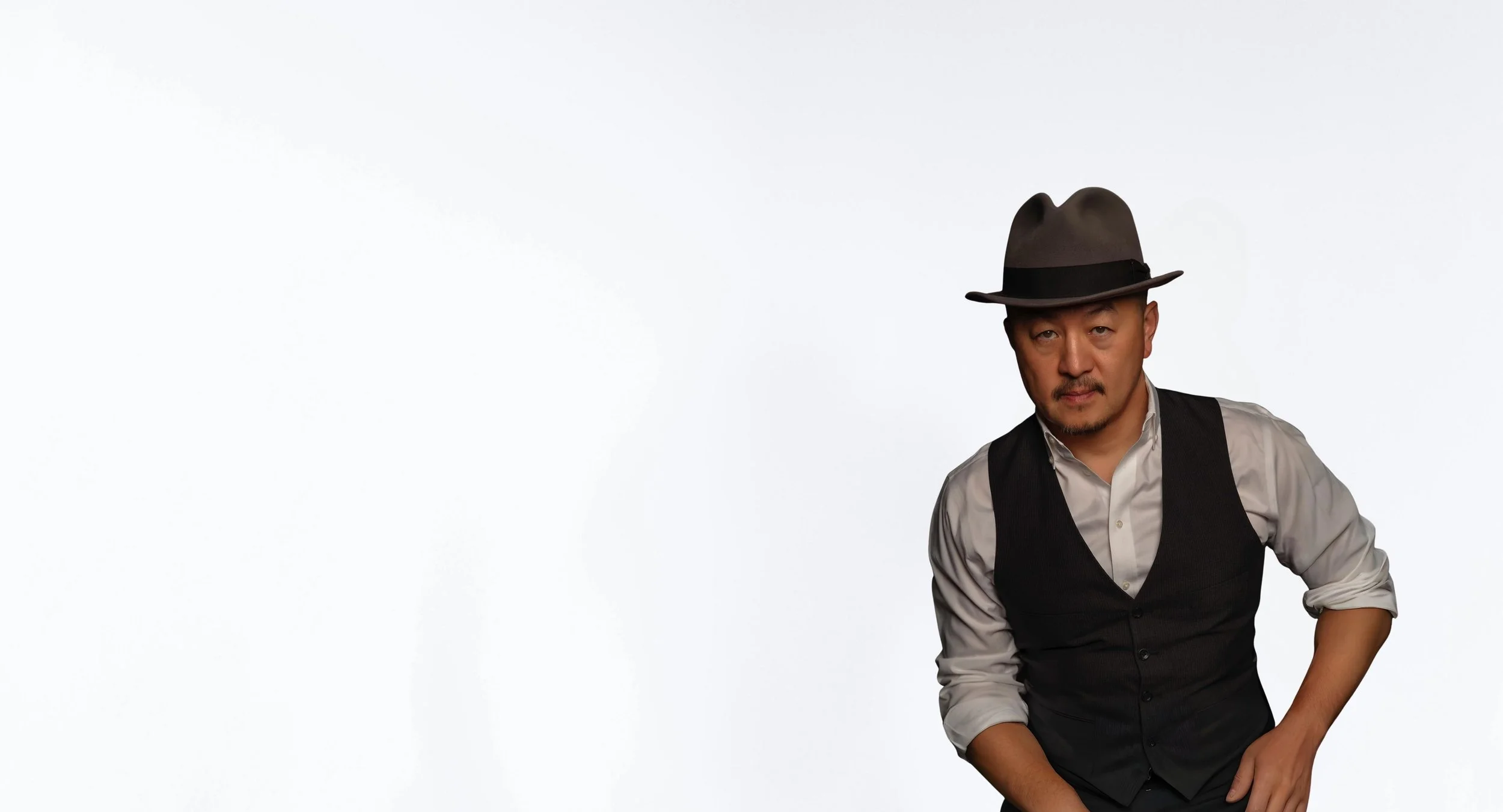 A man wearing a stylish fedora hat, a white dress shirt with rolled-up sleeves, and a black vest, standing against a plain white background