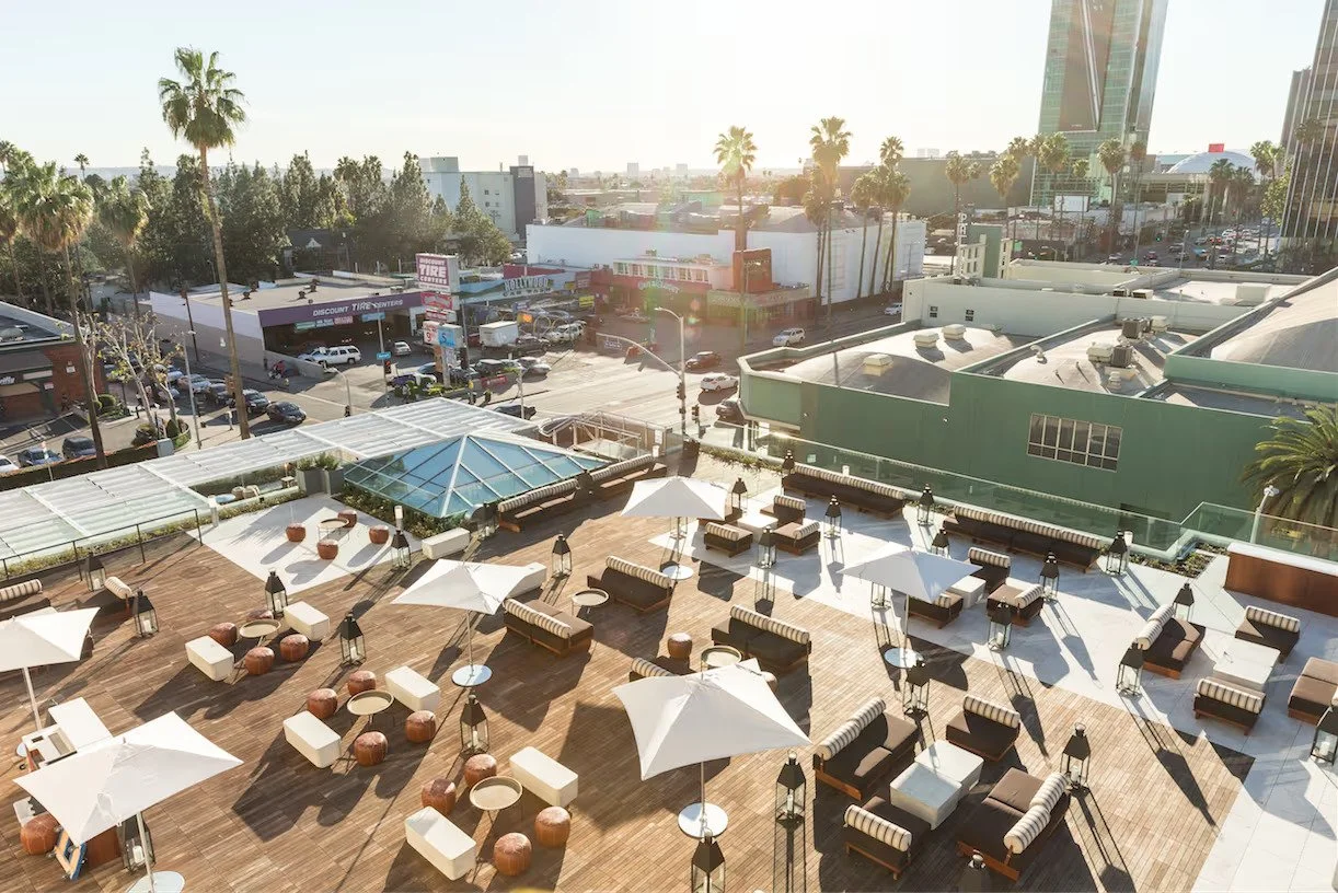 Rooftop lounge area with umbrellas, lounge chairs, and small tables, overlooking a cityscape with palm trees, buildings, and parking lot in the background during daytime.
