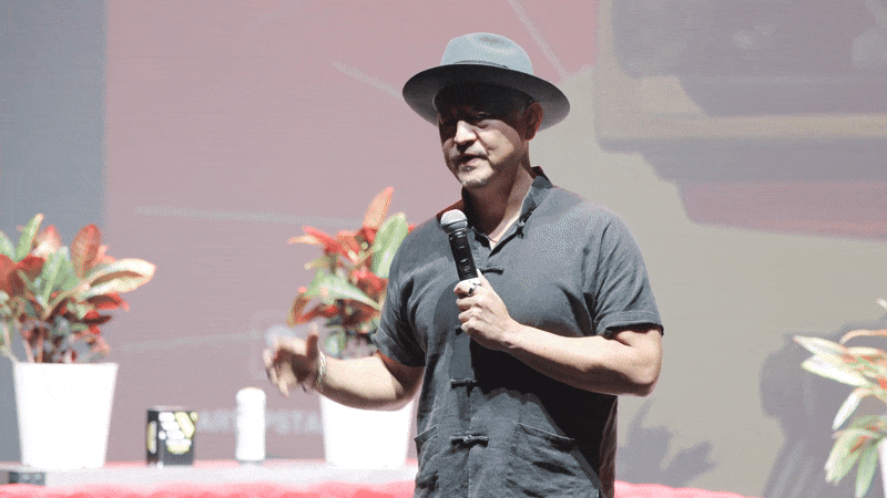 A man wearing a gray shirt and gray hat holding a microphone, speaking on stage with potted plants in the background.