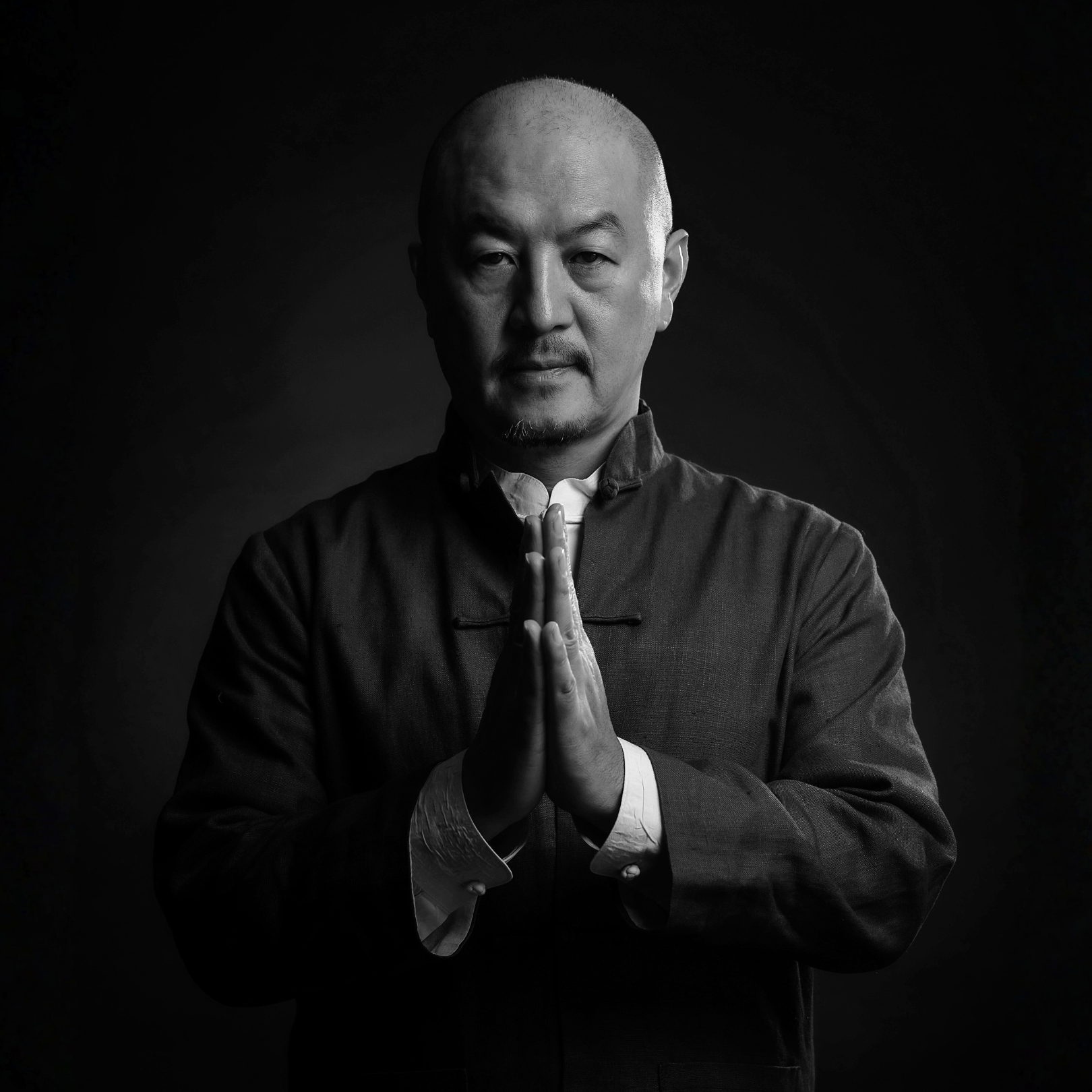 Black and white portrait of a man with a shaved head, wearing traditional Asian attire, with hands pressed together in a prayer-like gesture, looking directly at the camera.