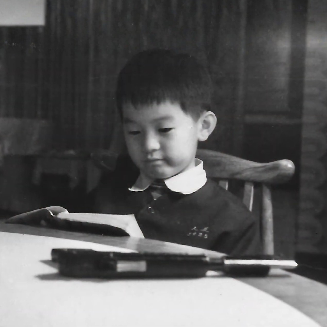 A young boy sitting at a desk, looking at an open book, with a pencil case on the desk in front of him, in black and white photo.