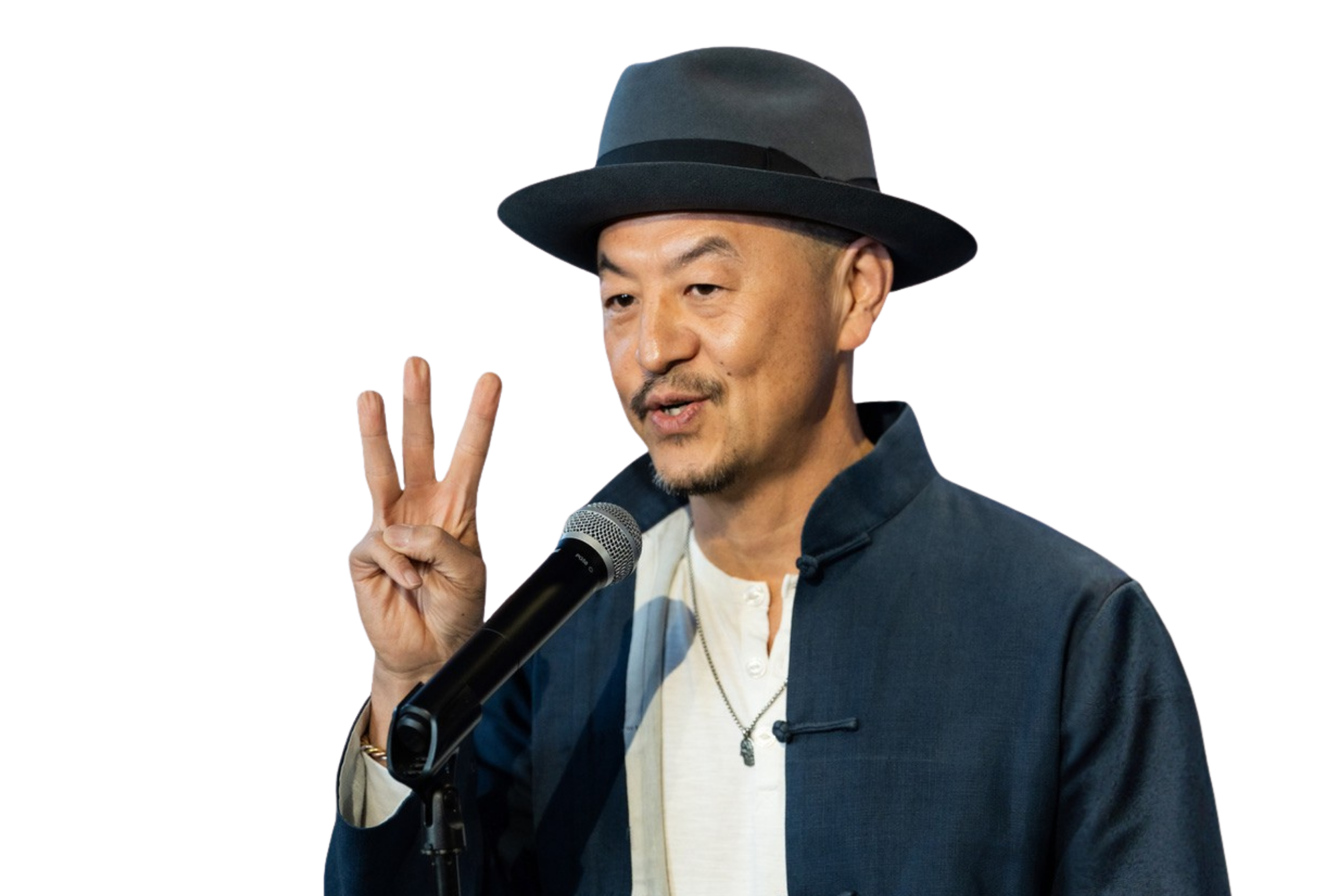 Man wearing a dark fedora hat, dark jacket over a white shirt, standing in front of a microphone, making a peace sign with his right hand, speaking or smiling.