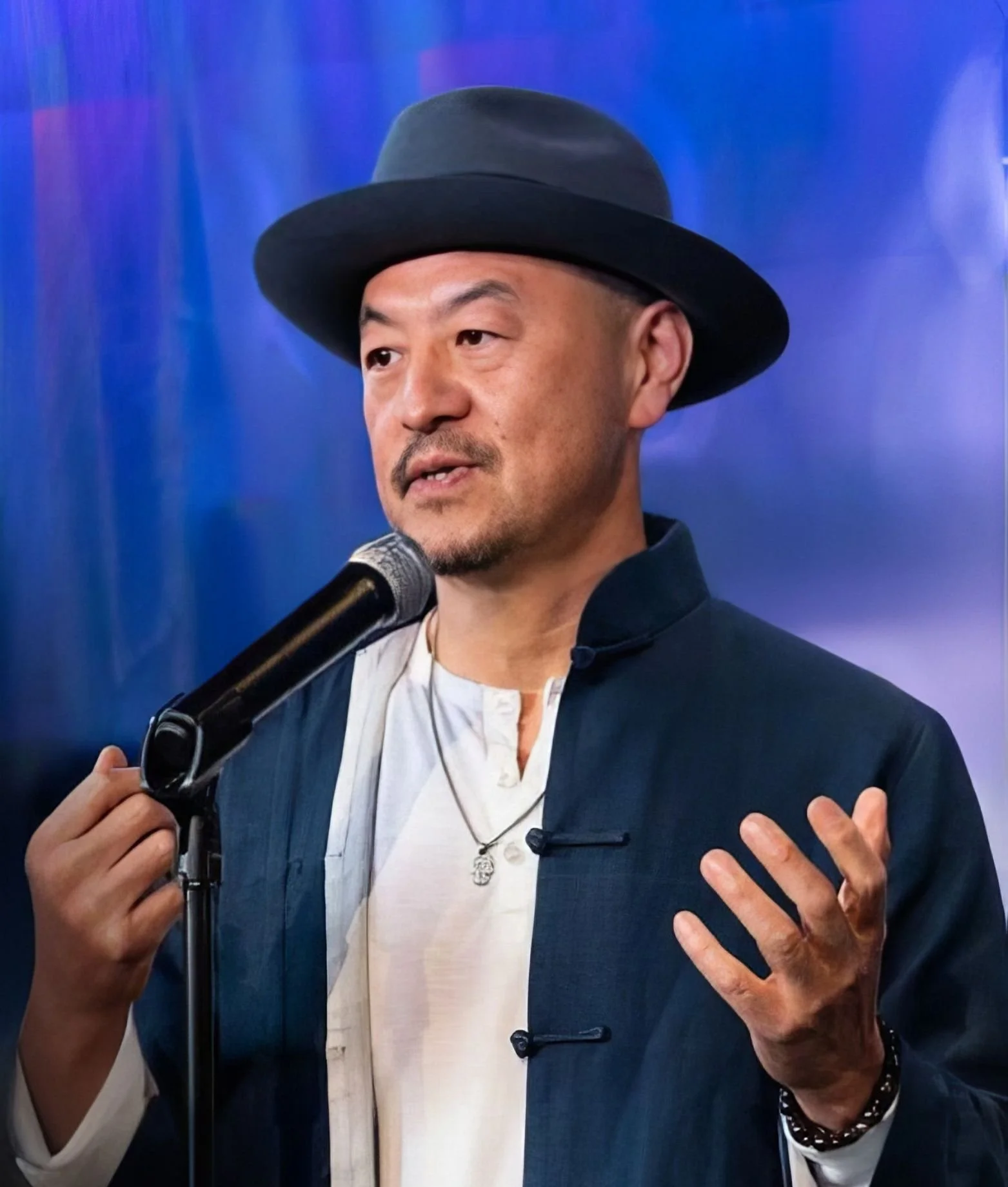 A man wearing a black hat and a dark blue jacket speaking into a microphone, with a background of purple and blue hues.