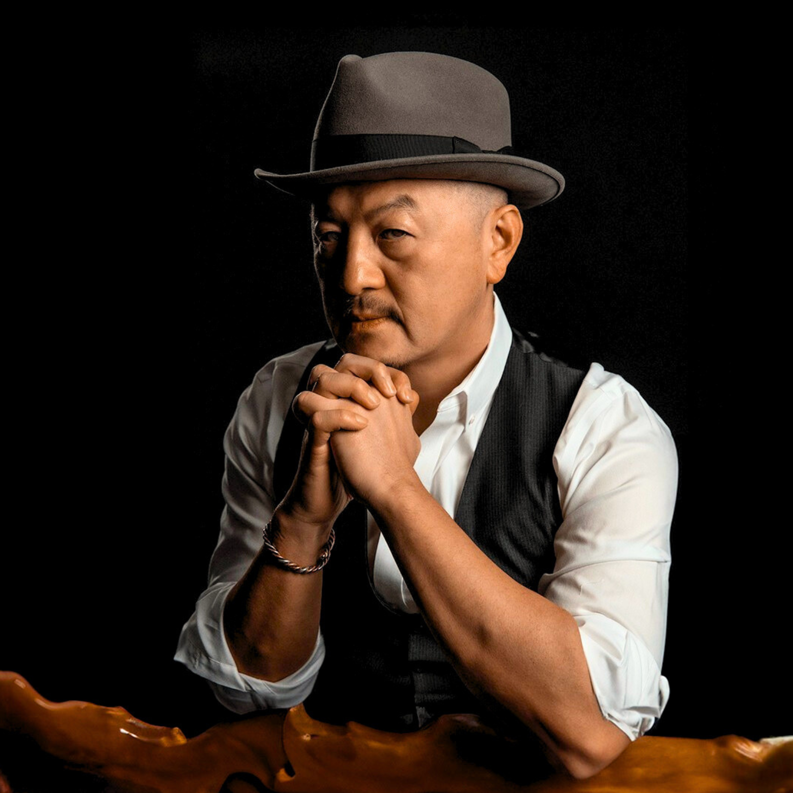 A man wearing a gray fedora hat, white shirt, and black vest, with hands clasped together near his face, against a dark background.