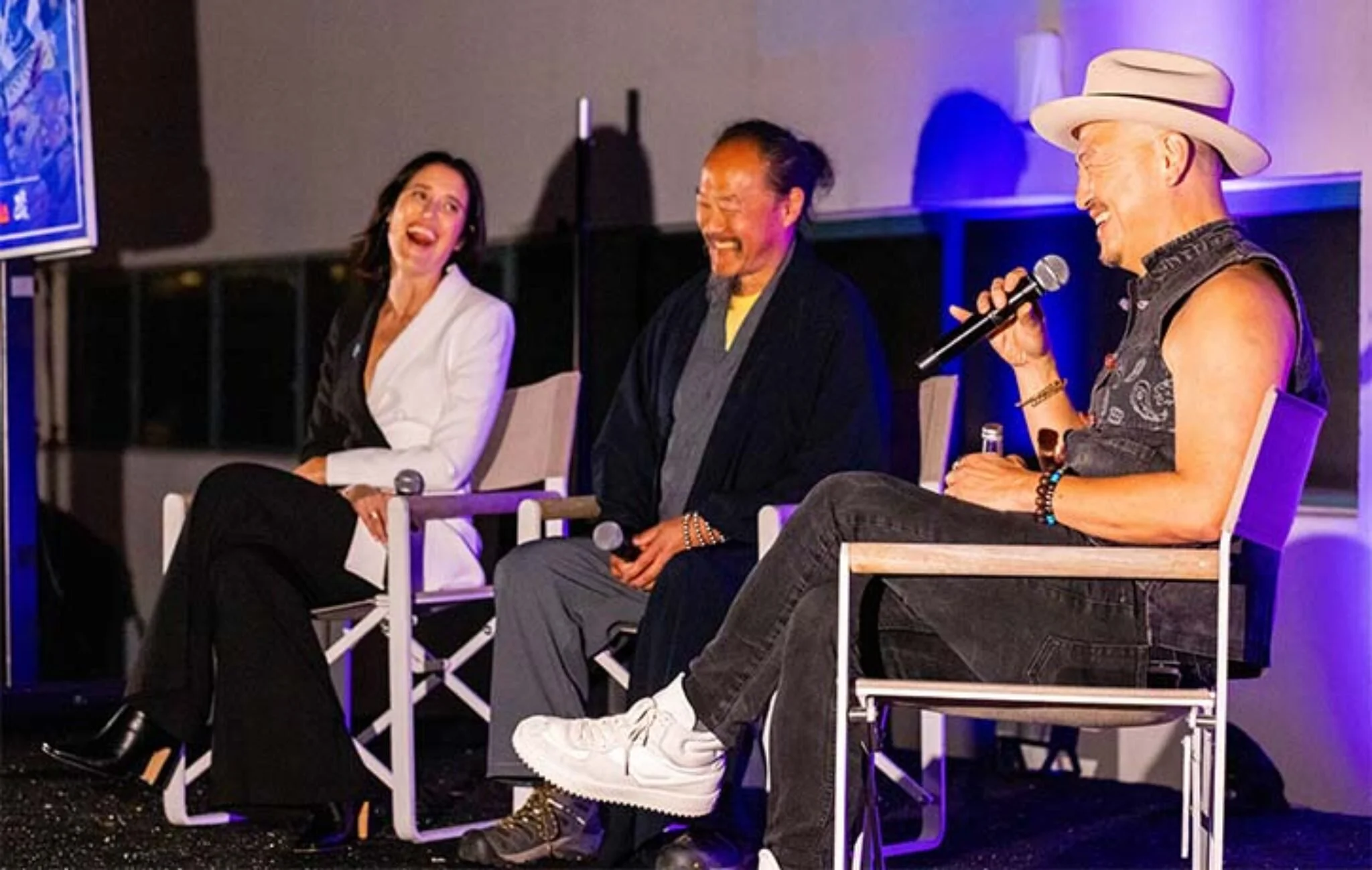 Three people sitting on stage, smiling and engaging in conversation. The woman on the left is wearing a white blazer and black pants. The man in the middle is dressed in traditional Japanese attire. The man on the right is wearing a sleeveless shirt, jeans, a hat, and holding a microphone.