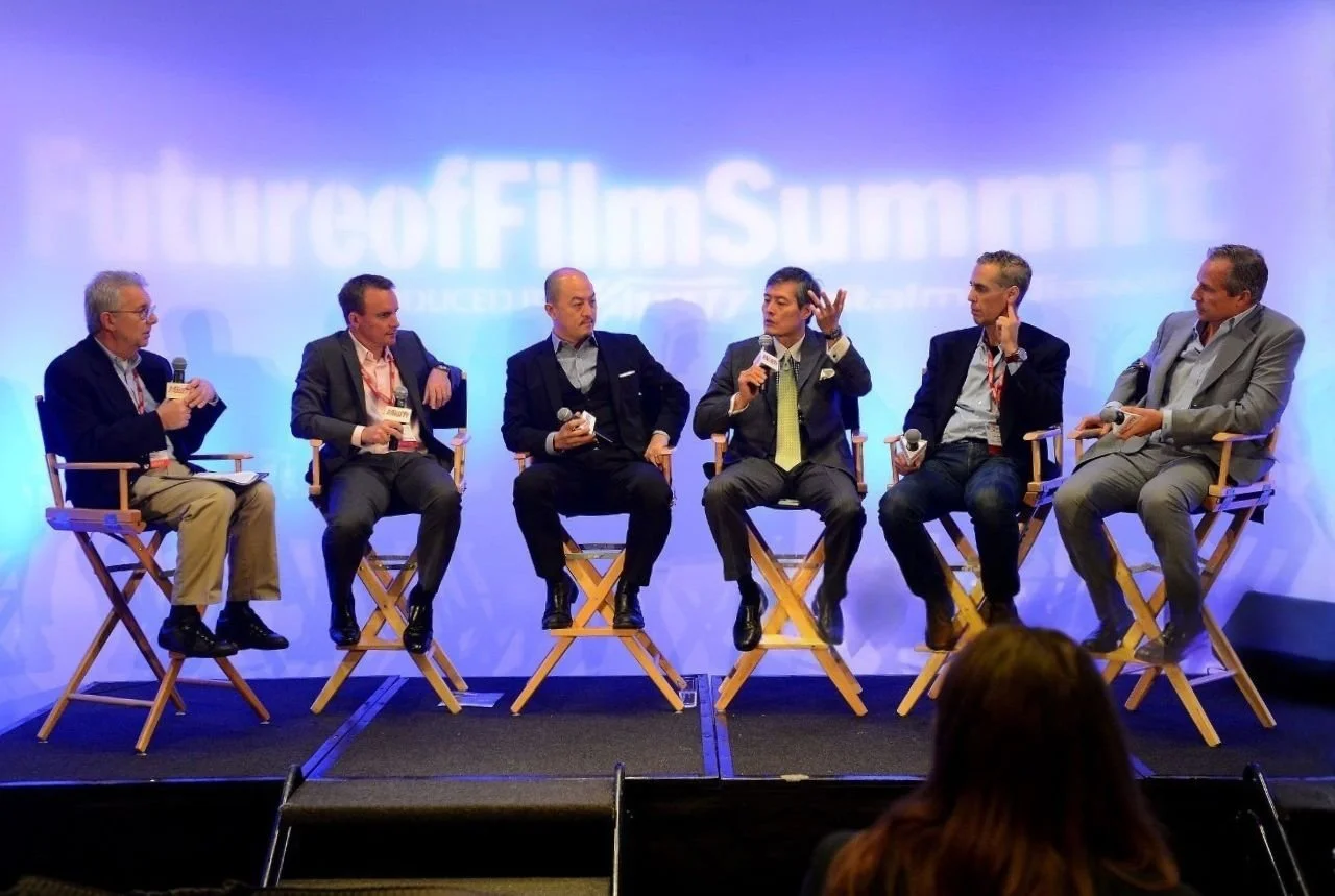 Six men sitting on stage in director's style chairs, participating in a panel discussion at the FilterCo Film Summer event, with a blue background and audience member in the foreground.
