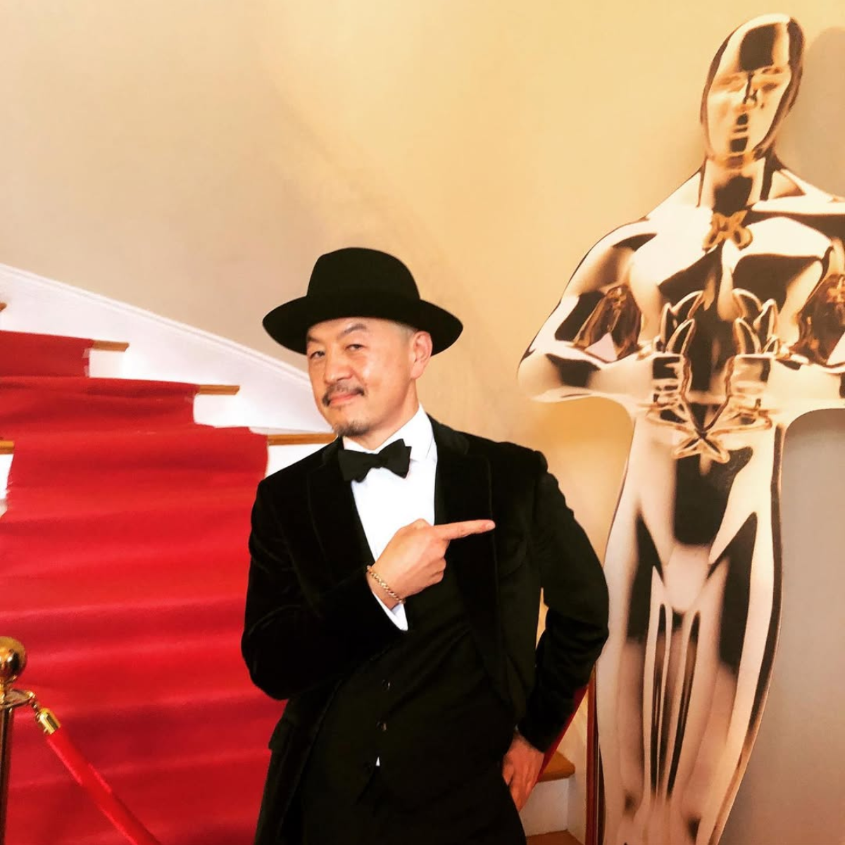 Man in tuxedo and bowtie pointing at an abstract female sculpture on a beige wall.