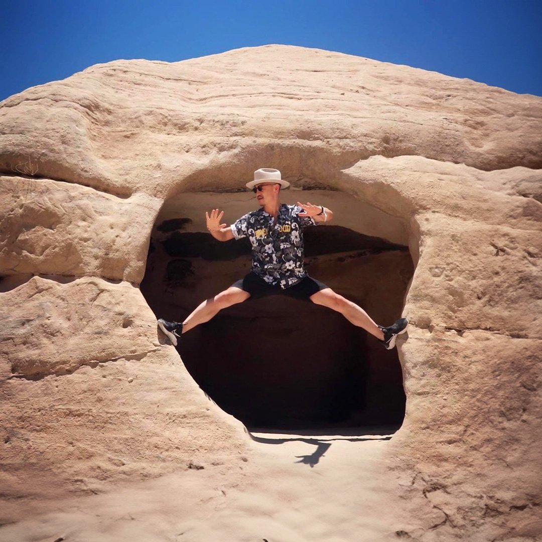 Person wearing a floral shirt, white hat, and sunglasses jumping with legs spread wide and arms raised in a rock formation with a large hole, against a bright blue sky.
