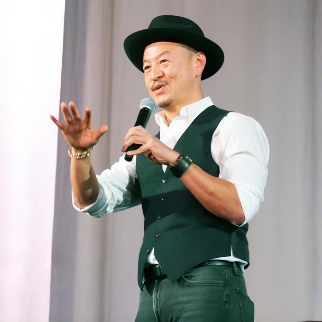 A man wearing a black hat, white shirt, and black vest, speaking into a microphone on stage.