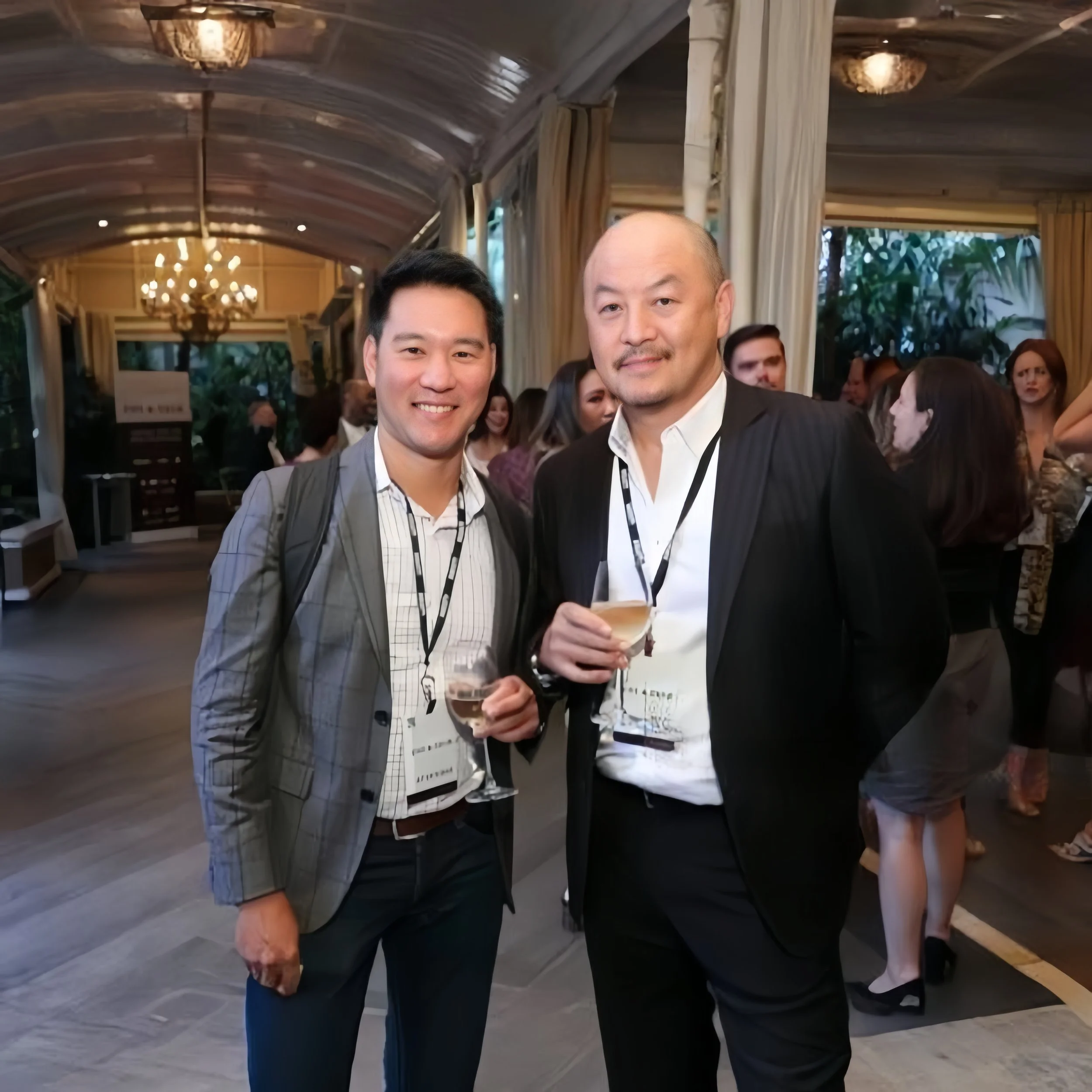 Two men at a social event, holding drinks, posing for a photo in a well-lit indoor venue with chandeliers and other people in the background.
