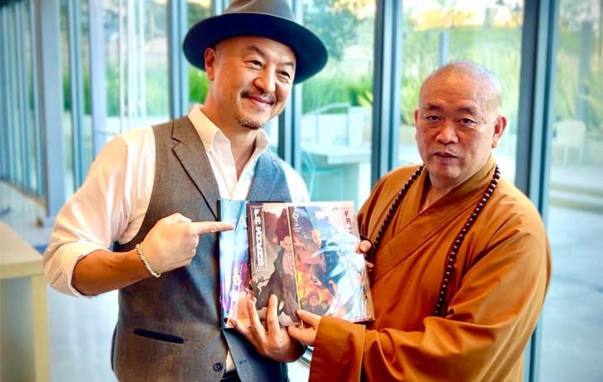 A man in a gray vest and white shirt, wearing a wide-brimmed hat, is smiling and pointing to a graphic novel held by a Buddhist monk in orange robes with a wooden bead necklace. The background features large glass windows revealing a lush, green outdoor area.