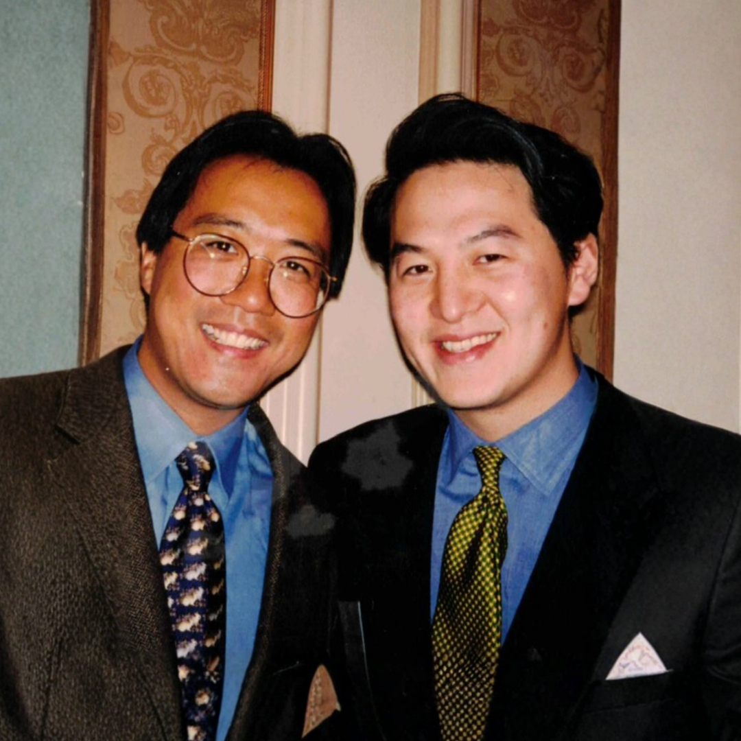 Two men smiling and dressed in suits and ties, posing indoors against a patterned wall.