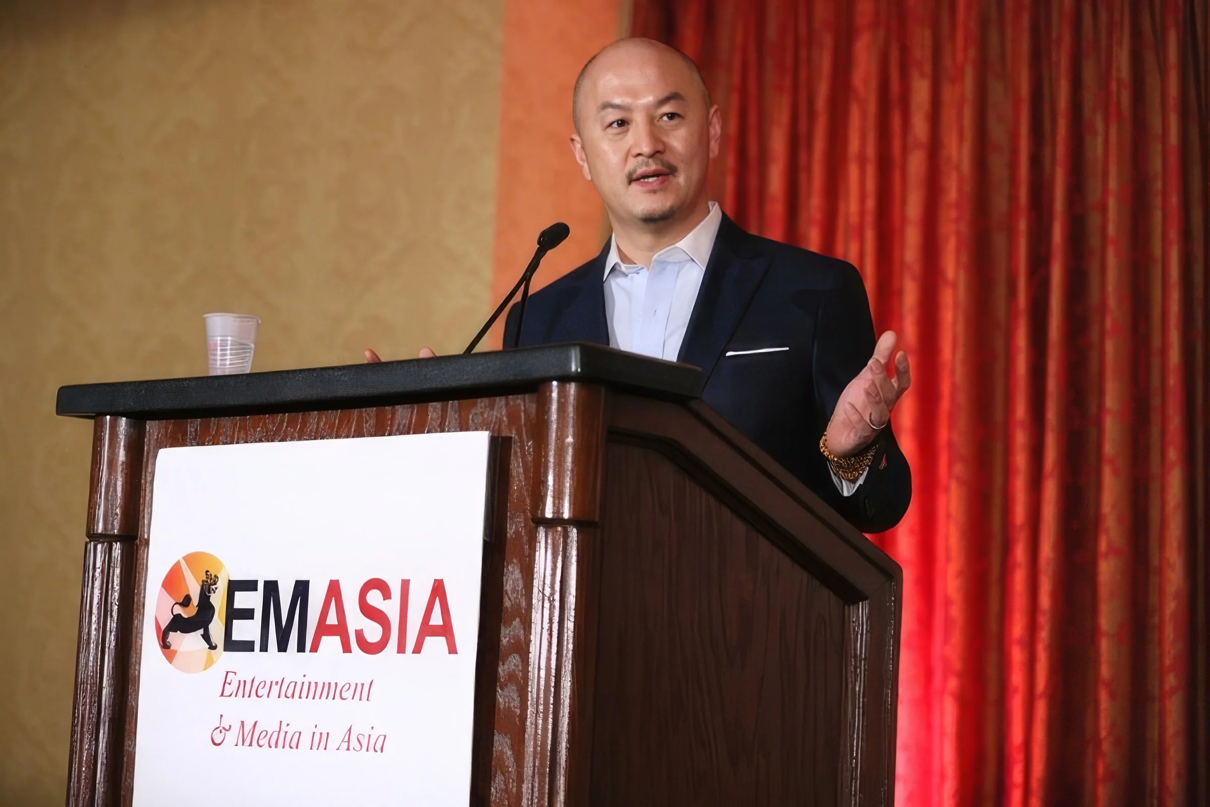 A man in a dark suit and white shirt speaking at a conference podium with a sign that reads "EMASIA Entertainment & Media in Asia," in front of an orange curtain.