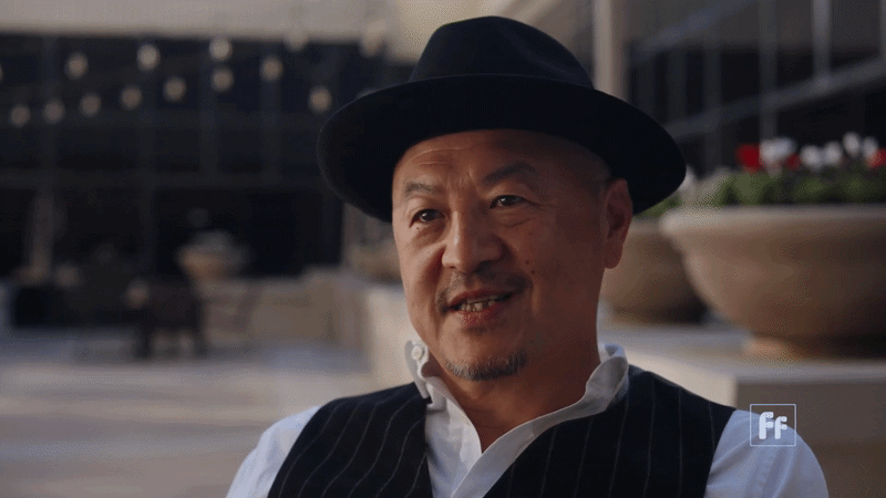 A man wearing a black hat and a pinstripe vest seated outdoors, smiling with a blurred urban background.
