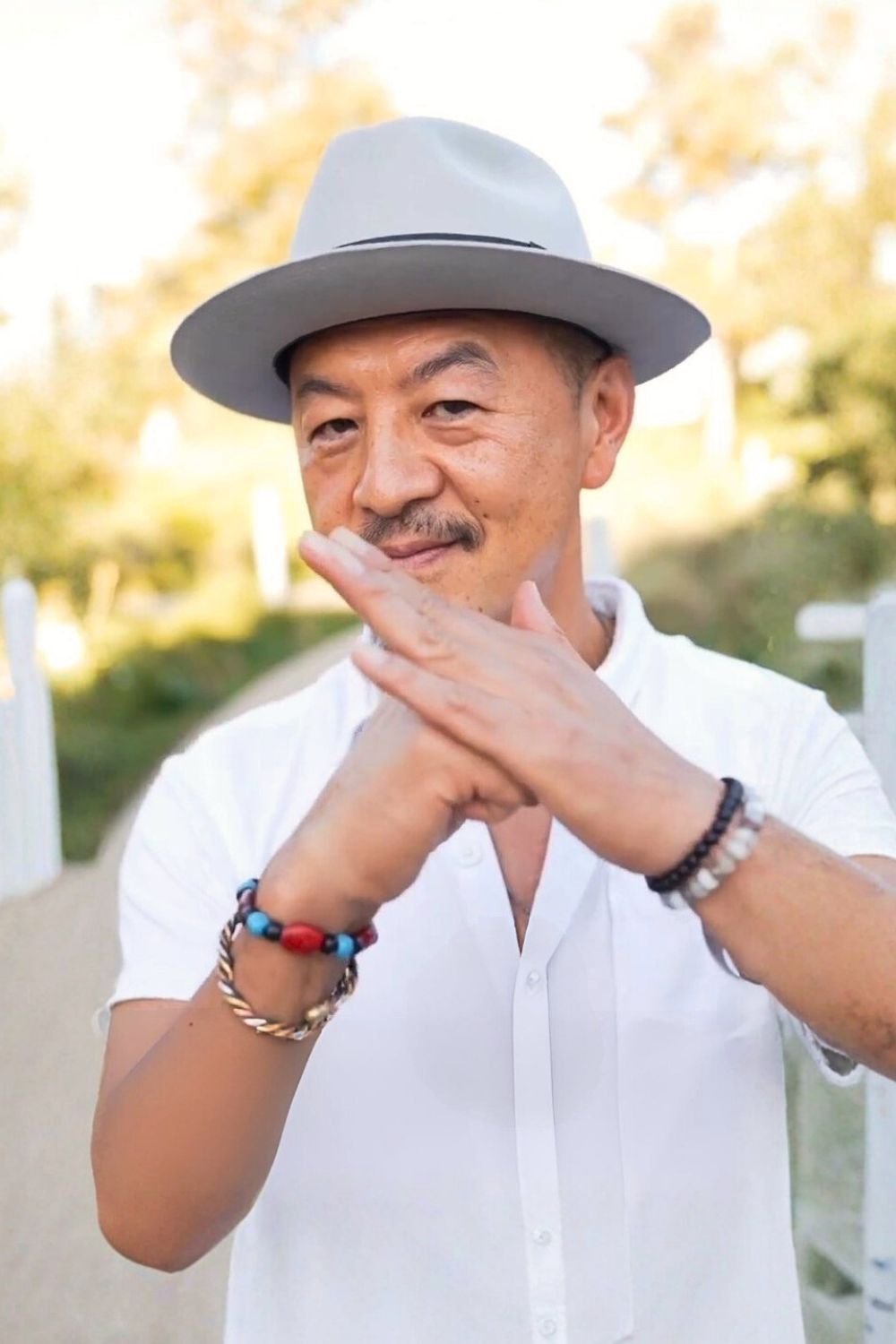 A man wearing a light gray fedora hat, white shirt, and bracelets on his wrists, posing outdoors with a warm smile, in a natural setting with blurry trees and sunlight in the background.