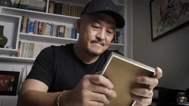 A man wearing a black cap and black t-shirt is sitting at a desk, holding and reading a book in a room with a bookshelf filled with books in the background.