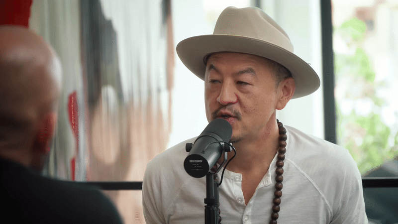 A man wearing a light-colored hat and white shirt speaking into a microphone during a recorded interview or podcast, with another person partially visible in the foreground.