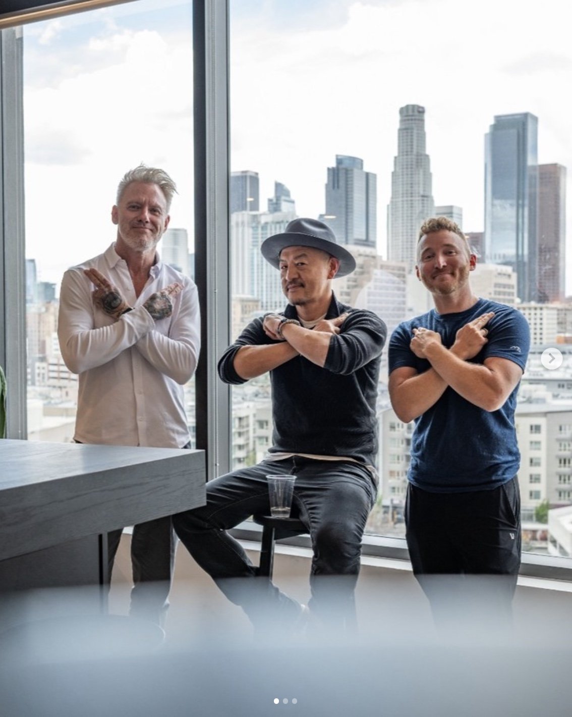 Three men posing with arms crossed in front of a city skyline window