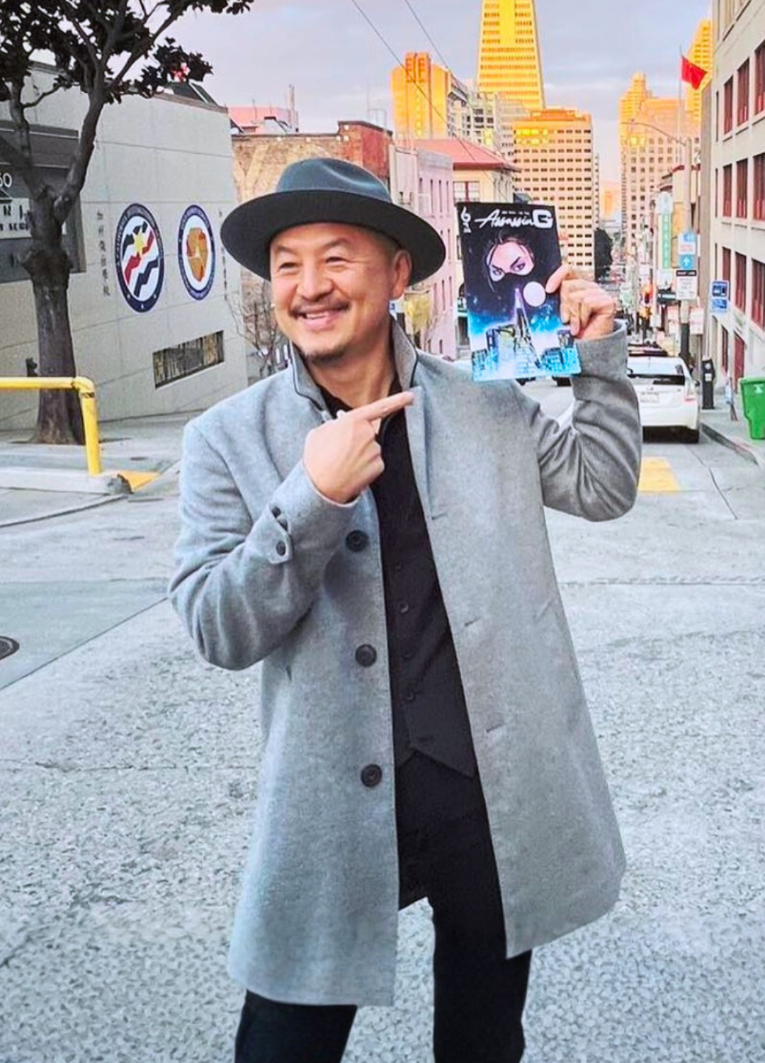 A man smiling in an urban street, wearing a gray coat and a black hat, holding up a comic book titled "Aiirna" with cityscape artwork on the cover.