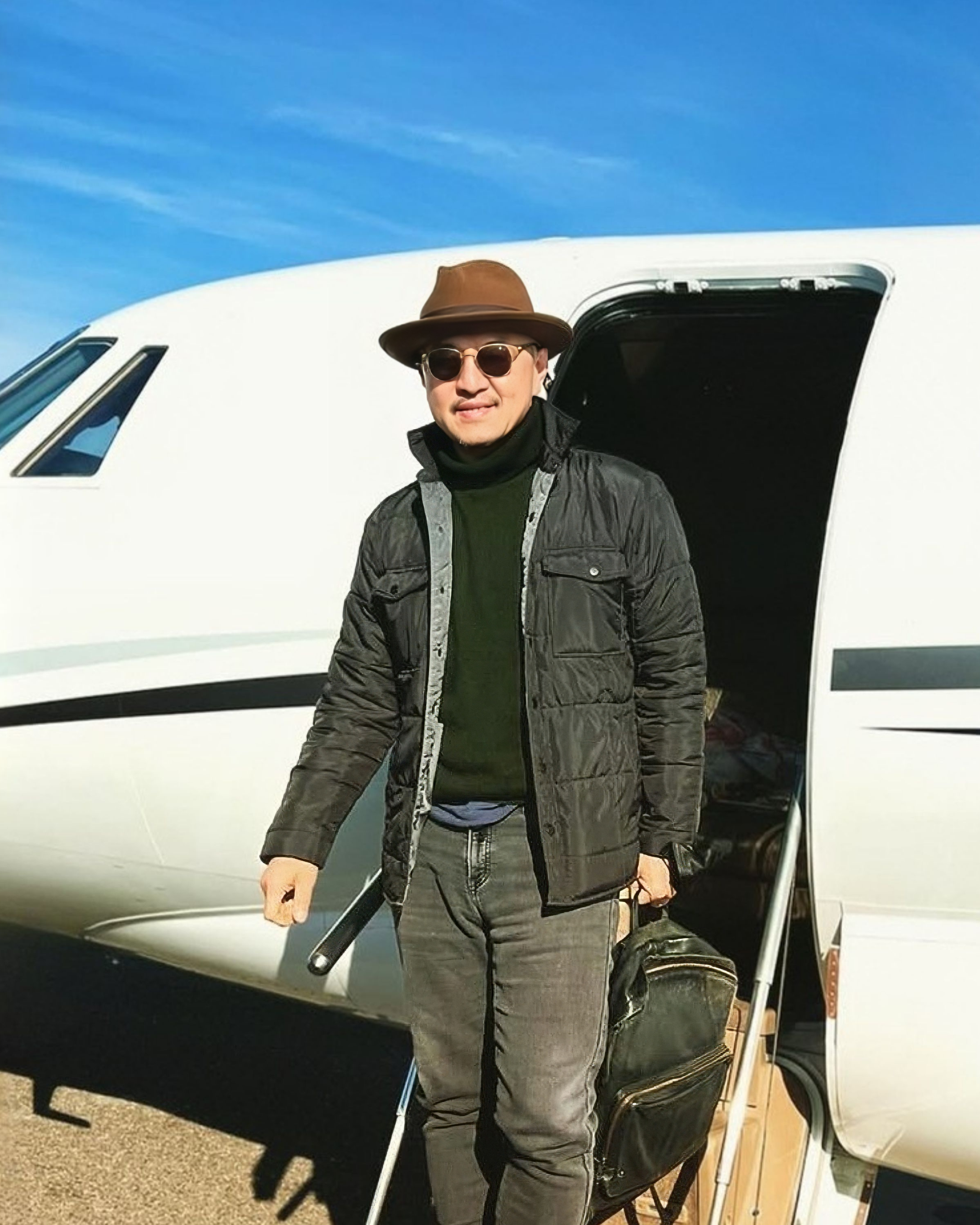 Man in sunglasses, brown hat, black jacket, green sweater, gray jeans, boarding private jet with baggage.