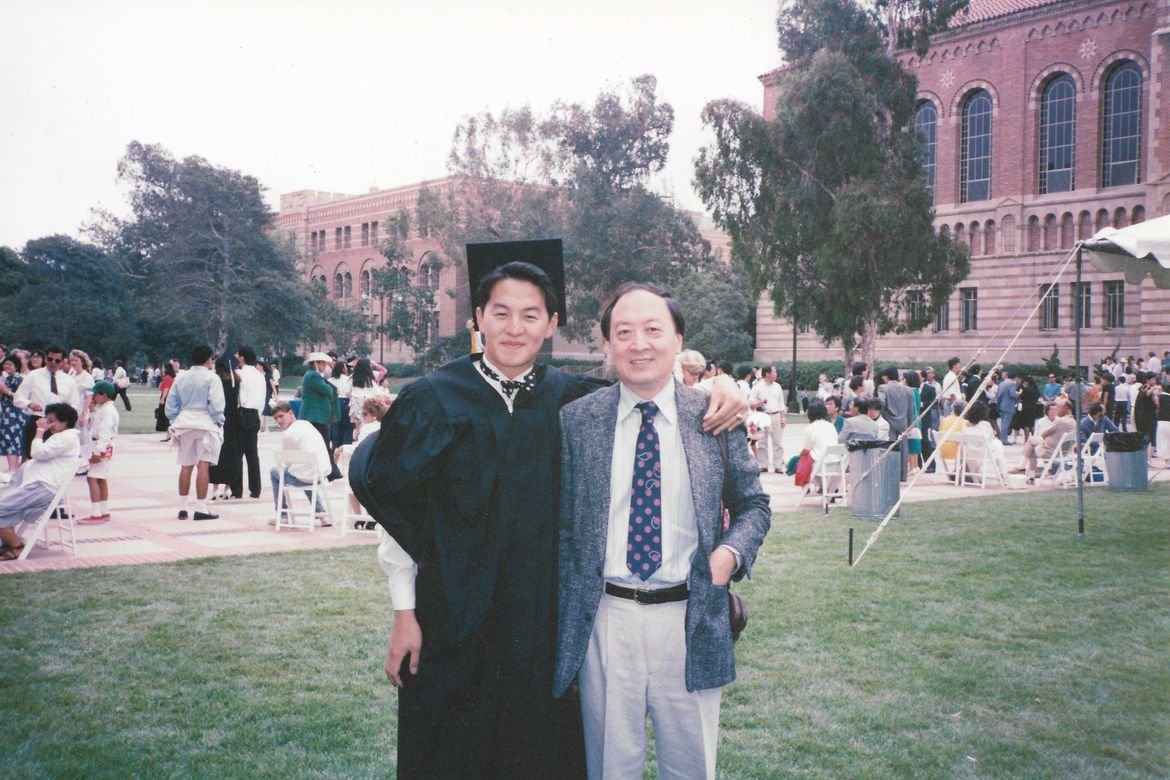 Two men smiling at a graduation ceremony outdoors, one in a graduation gown and cap, the other in a suit. People are seated and standing in the background with a large brick building and trees.