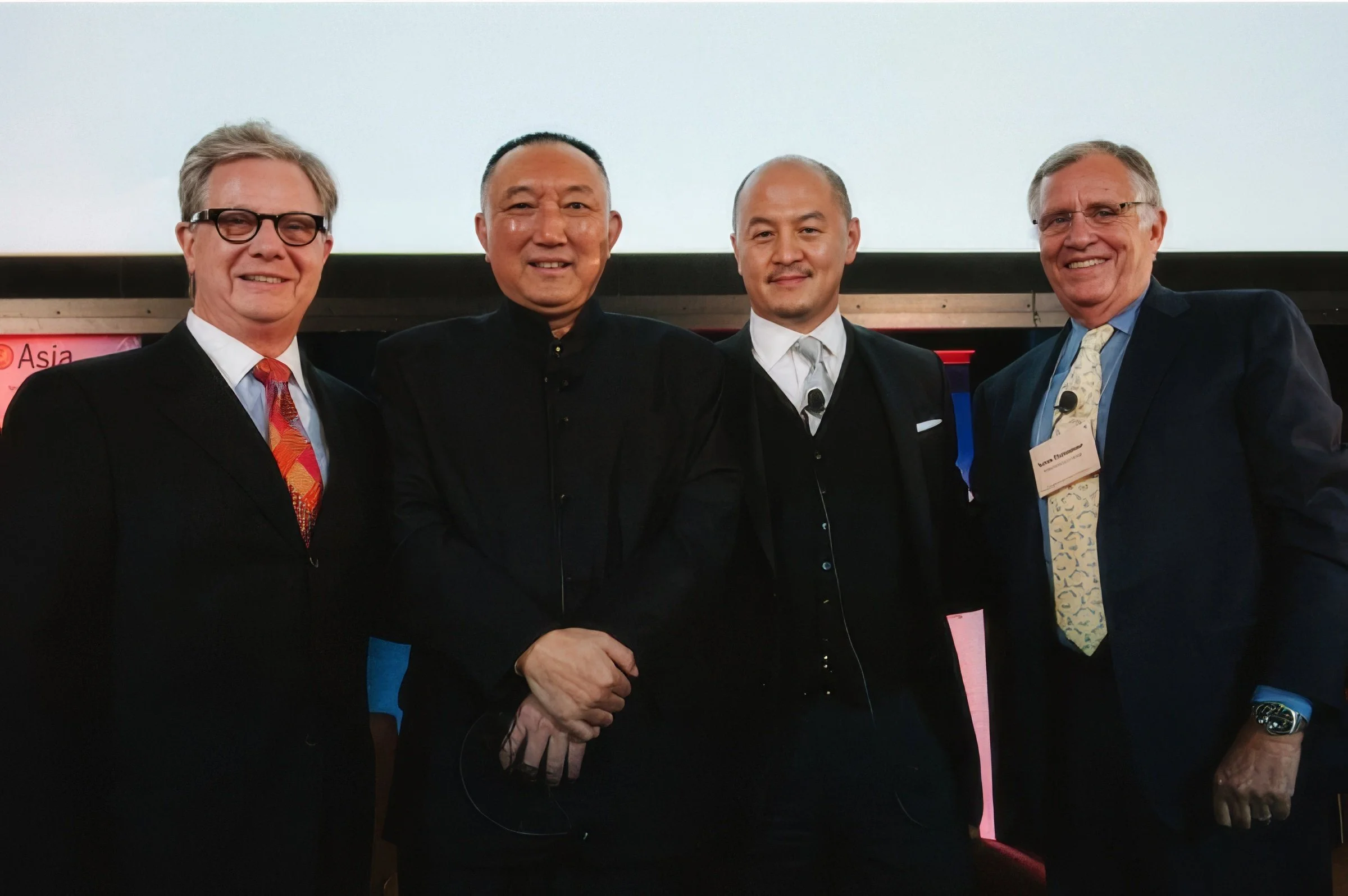 Four men in business suits standing together at an event, smiling at the camera.