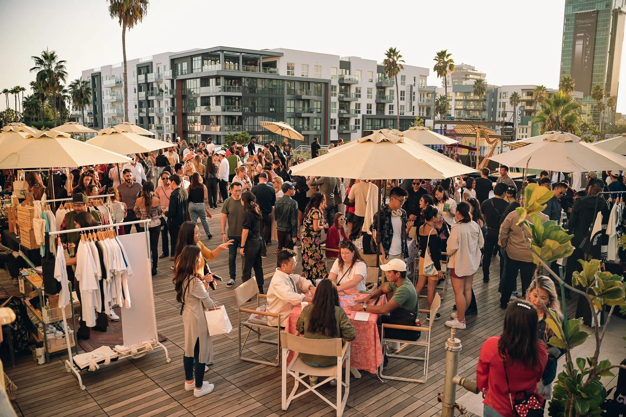A bustling outdoor market on a wooden deck with people shopping, socializing under beige umbrellas, trees, and modern buildings in the background.