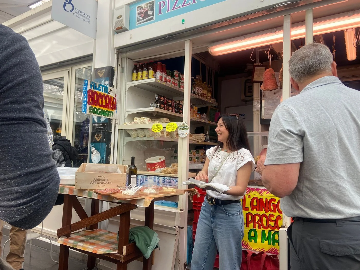 A woman smiling and talking to a man at an outdoor food stand, with tables, bottles, and various food items displayed.