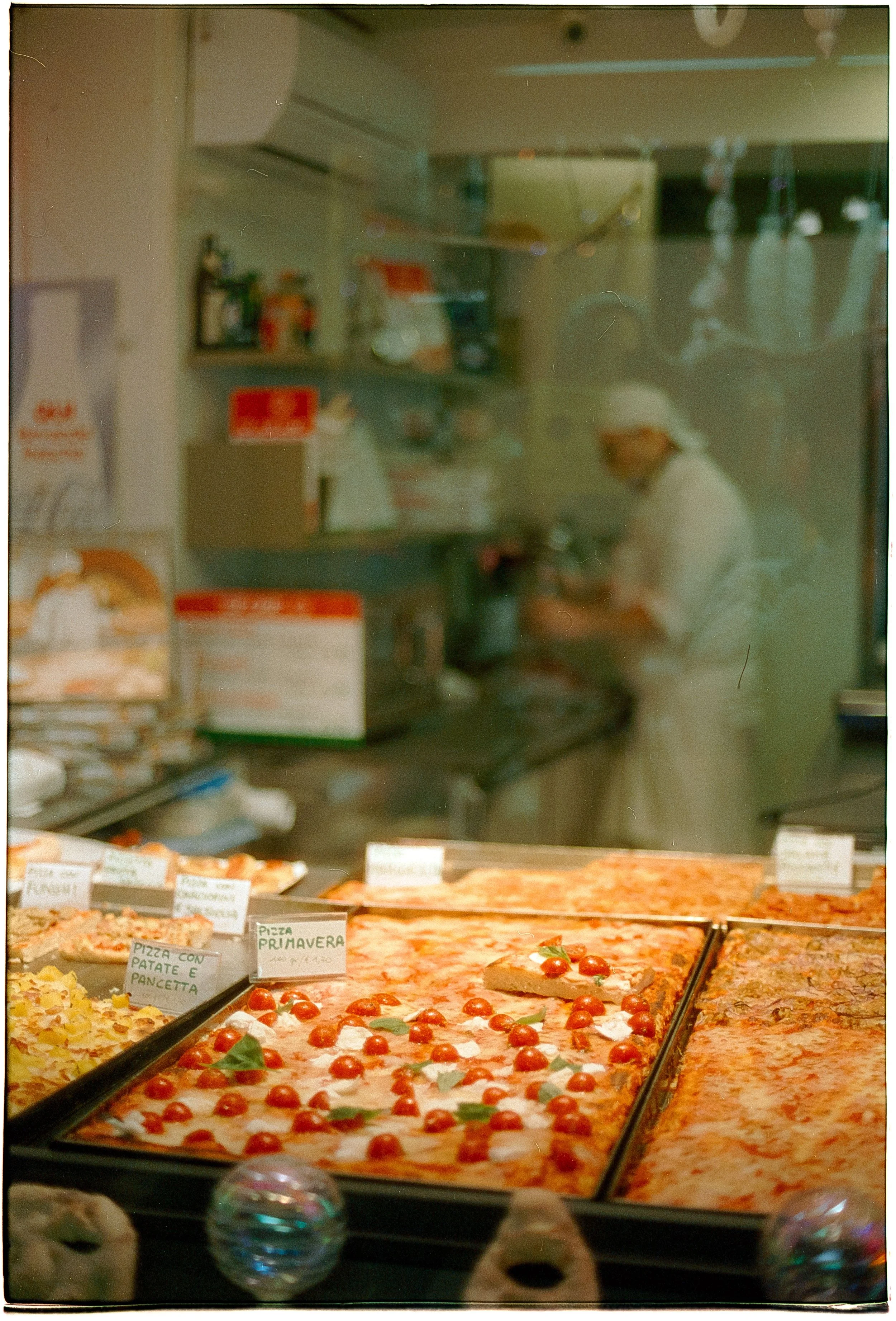 A pizza display case with various types of pizza, including one topped with cherry tomatoes. There are handwritten signs labeling the pizza types, such as 'Pizza Primavera' and 'Pizza con Patate e Pancetta'. A worker wearing a white apron and hairnet is behind the case preparing food in the background.