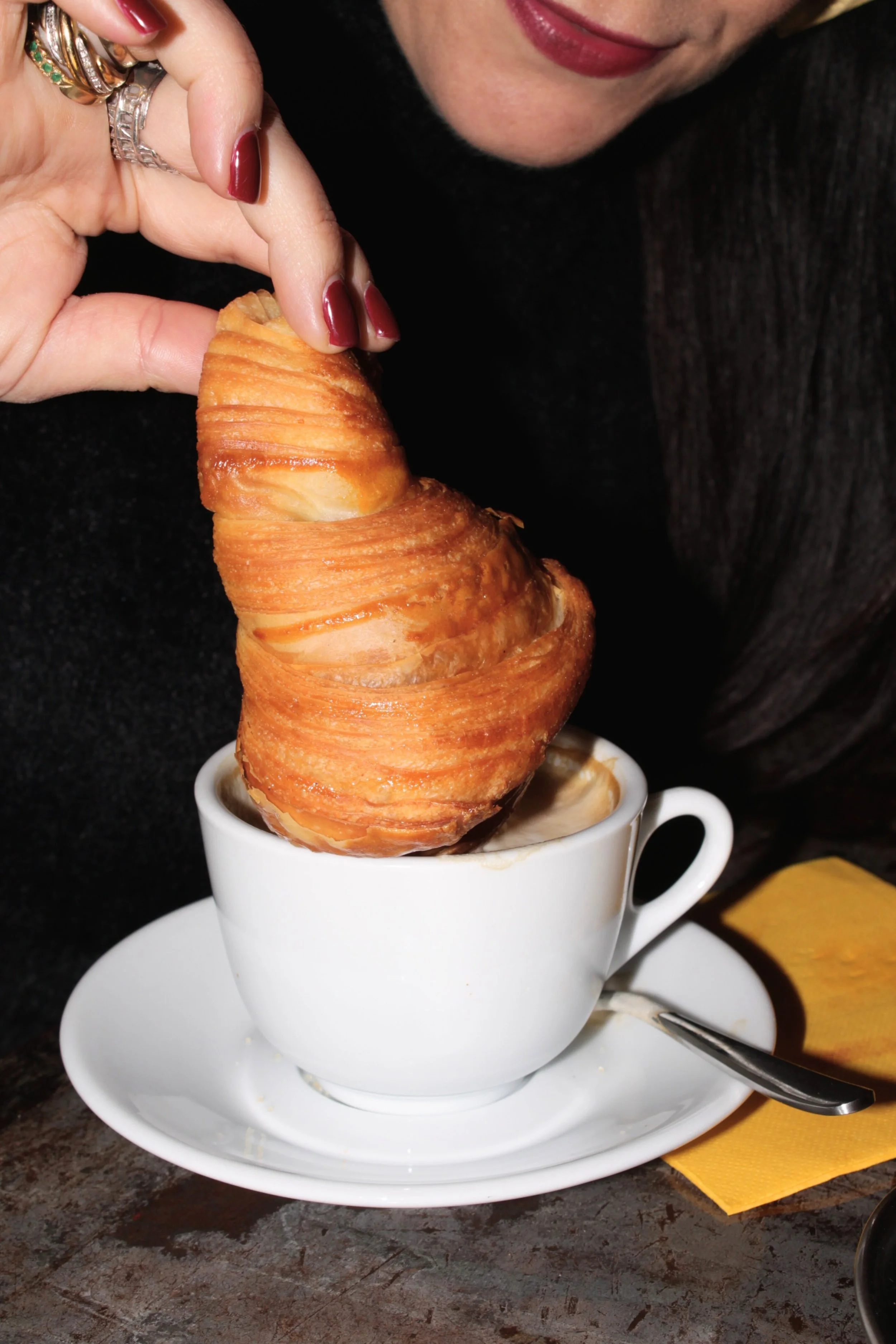 A woman with red lipstick and rings on her fingers is holding a croissant over a cup of coffee. The croissant is dipped into the coffee, and she is about to eat it.