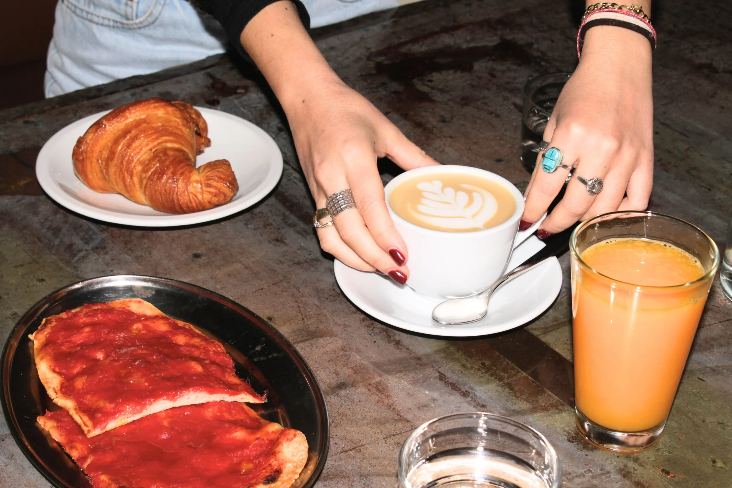 Person's hands with rings, holding a cup of coffee with latte art, on a table with a croissant, three slices of pizza, a glass of orange juice, and a glass of water.