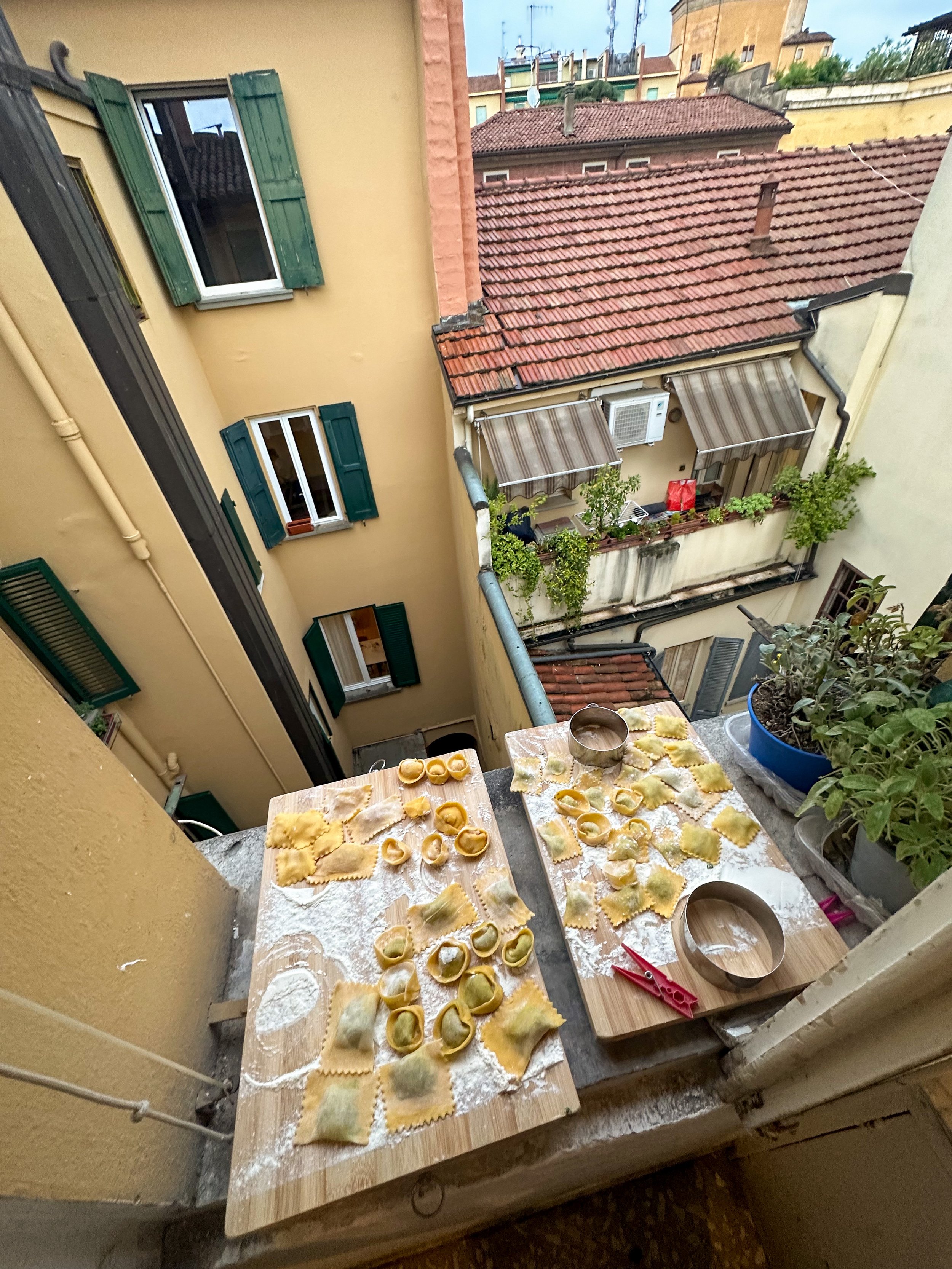 Two wooden tables on a balcony with homemade pasta ready to be cooked, surrounded by potted plants, with a cityscape of apartment buildings and rooftops in the background.