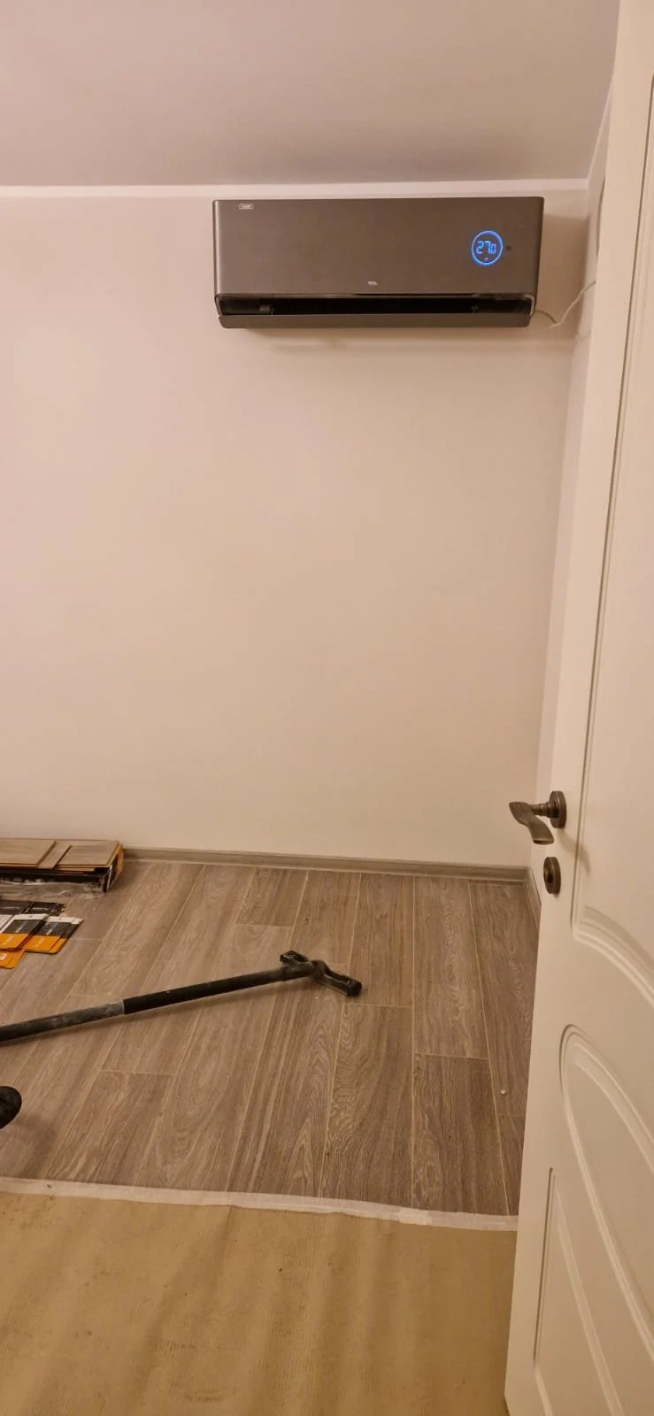 An unfinished room with a wall-mounted air conditioner displaying 27°C, a vacuum cleaner on the floor, and a door partially open.