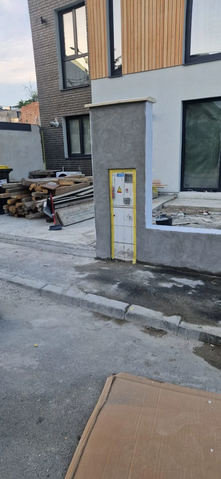 Construction site with a new building, stacks of wood, construction tools, and an electrical panel on a concrete wall near the sidewalk.