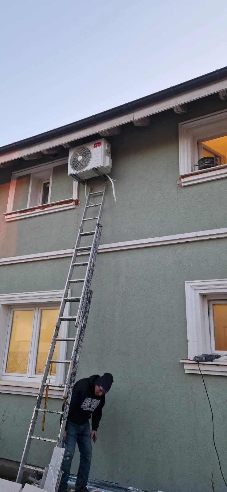 A person standing on the ground next to a ladder, working on the underside of an air conditioning unit mounted on the exterior wall of a house. The house has green walls, white-framed windows, and a black roof.
