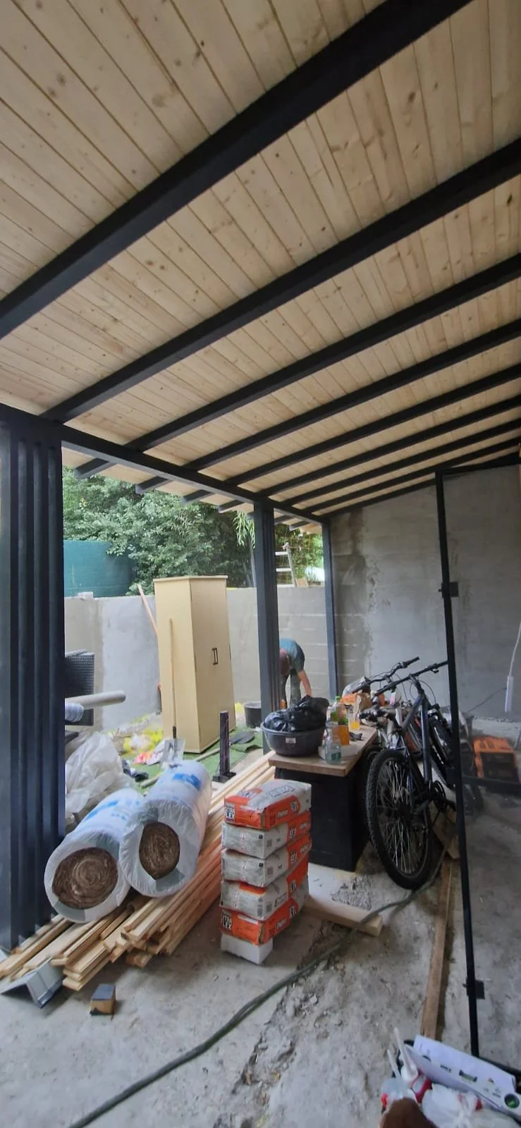 A covered outdoor space under construction, with building materials, a bicycle, and a person working in the background.
