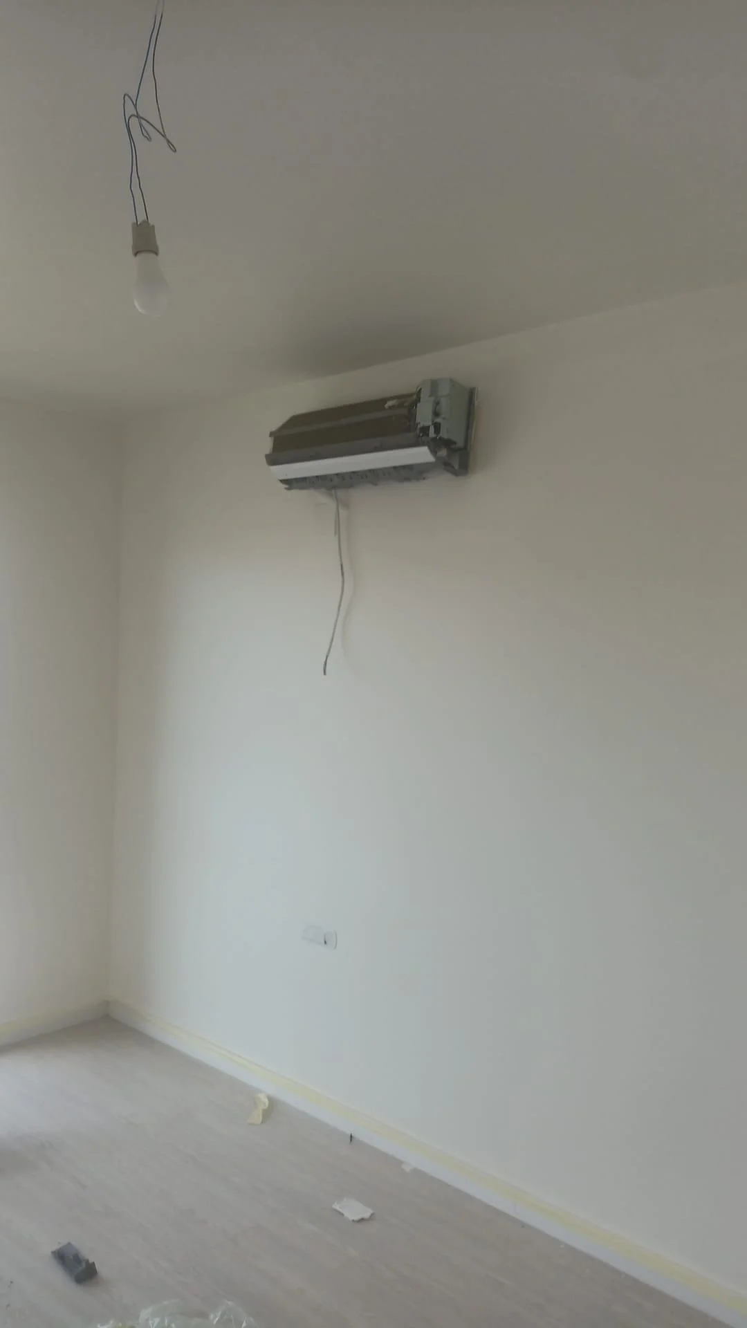 Empty room with an air conditioning unit installed on the wall, an exposed light bulb hanging from the ceiling, and some debris on the floor.