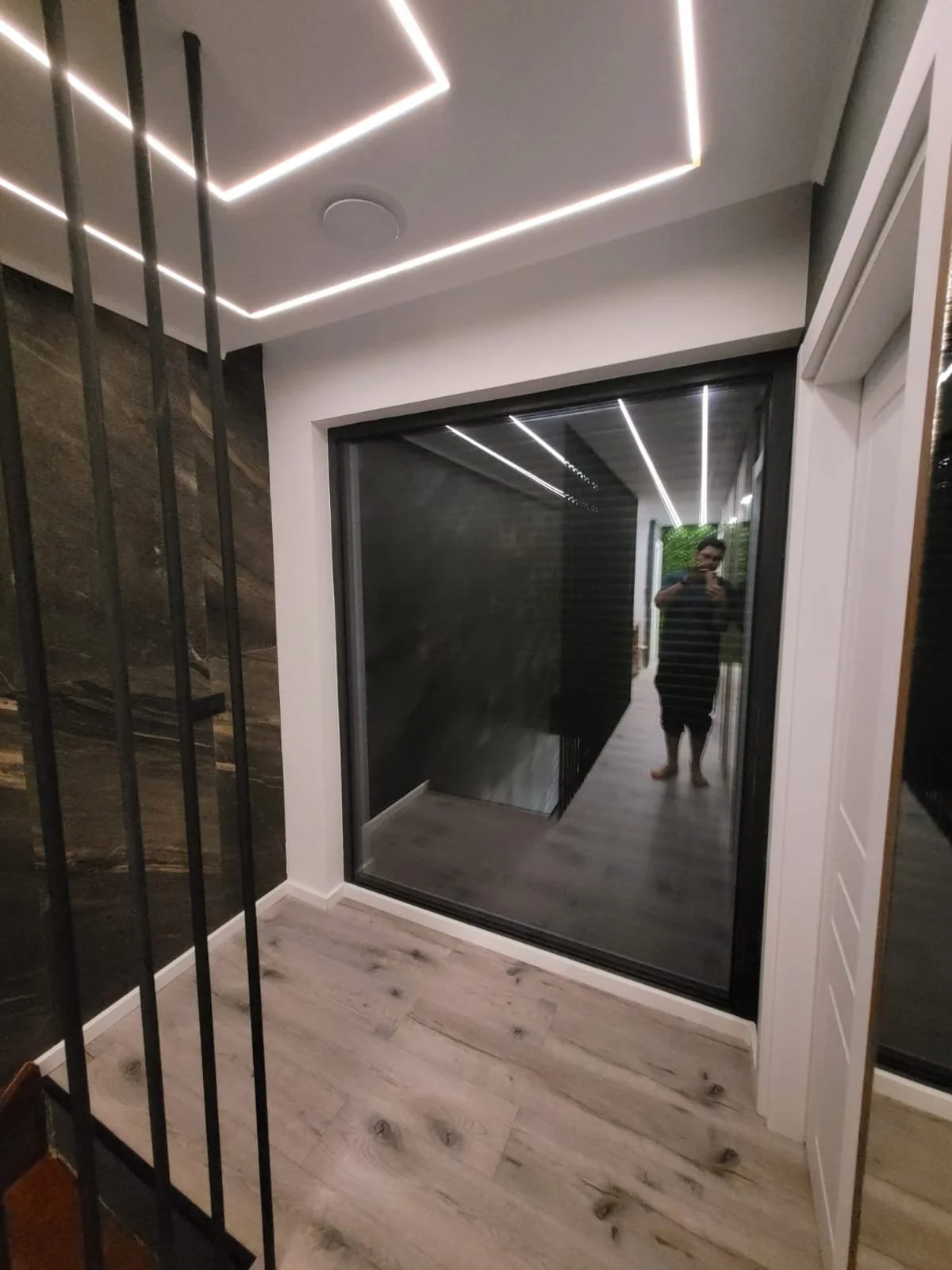 A modern hallway with a sliding glass door, wood flooring, and black metal bars on the left side. The ceiling features white LED strip lighting in geometric patterns. A person is taking a photo reflected in the glass.