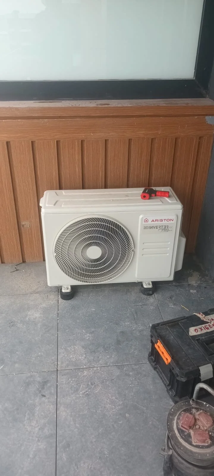 White Ariston air conditioning unit installed on a tiled floor in front of a wooden panel wall with a window above it.