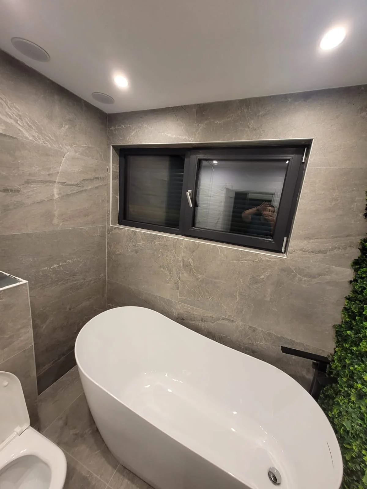 Modern bathroom with gray tiled walls, a white freestanding bathtub, a window with dark frames, and a partial view of a toilet on the lower left corner.
