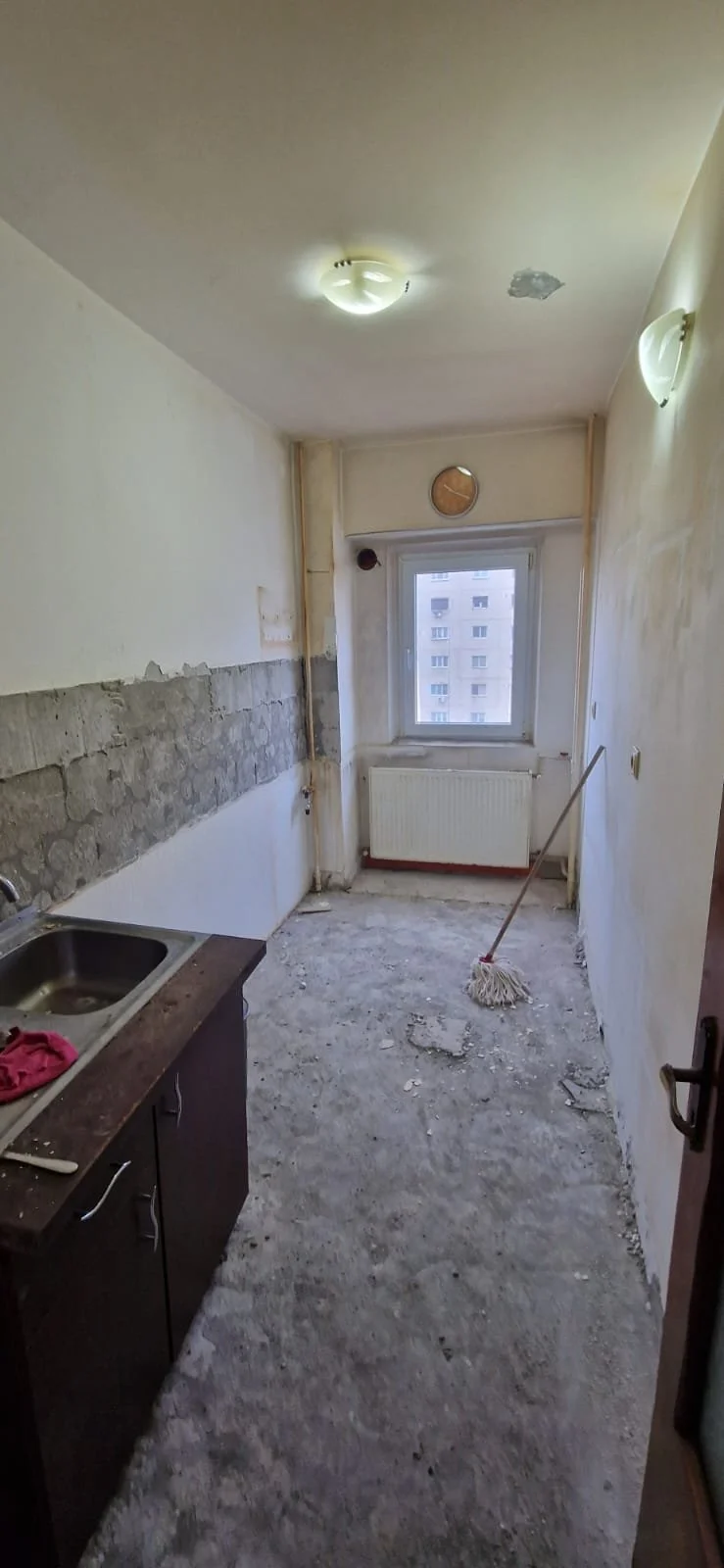 Empty, under-renovation kitchen with a window, a radiator, a mop on the floor, and unfinished walls.