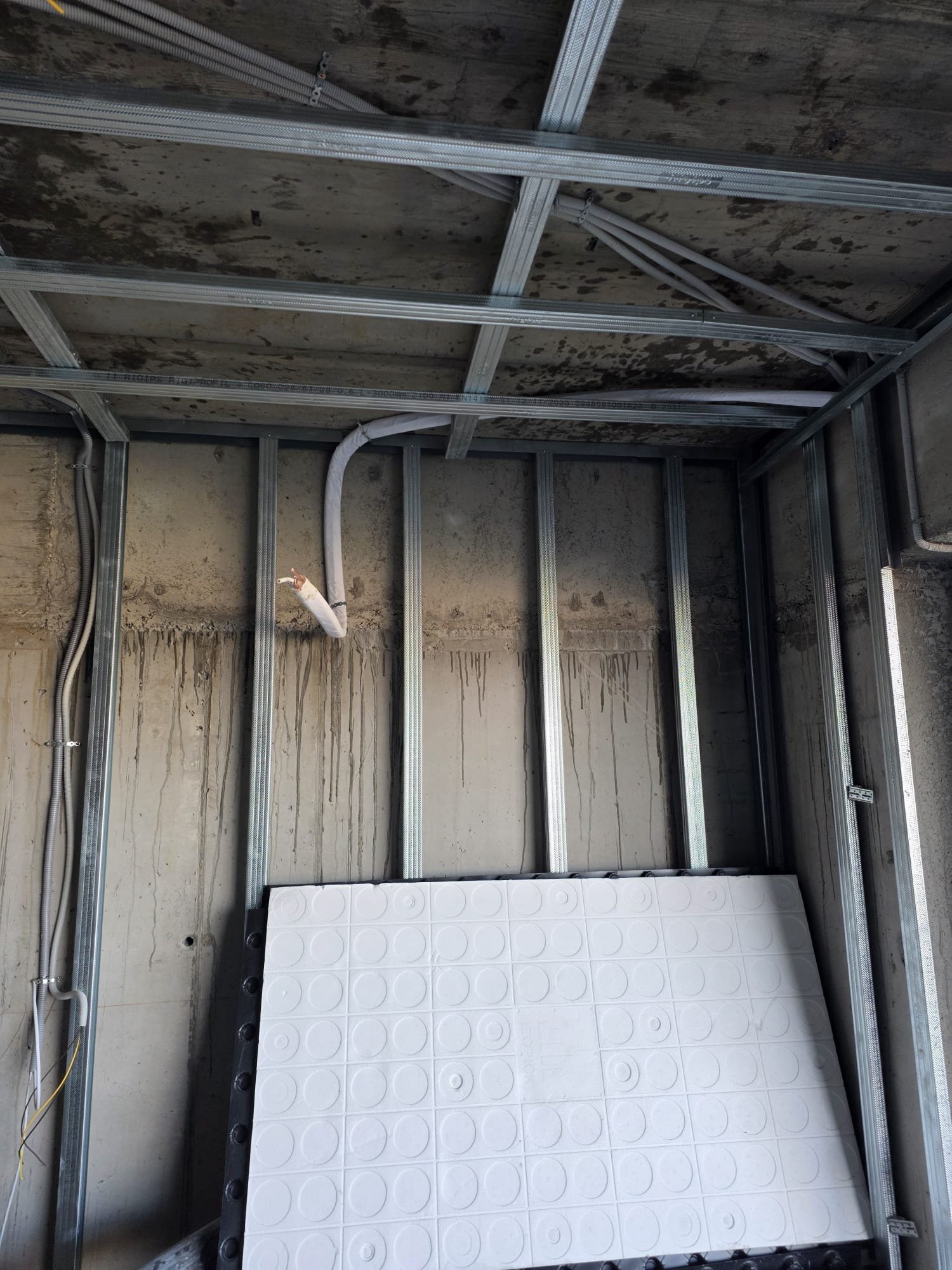 Interior view of a construction site with metal framing and exposed wiring, ceiling showing moisture stains, and a large white panel leaning against the wall.