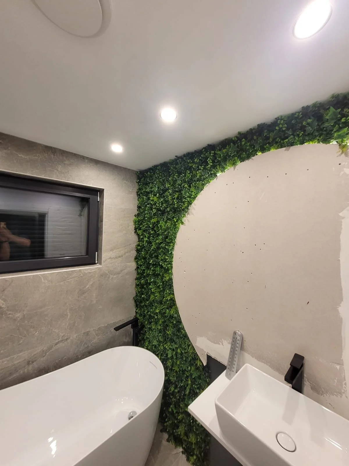Modern bathroom with grey textured wall, black-framed window, white bathtub, white vessel sink, and a decorative green leafy wall feature