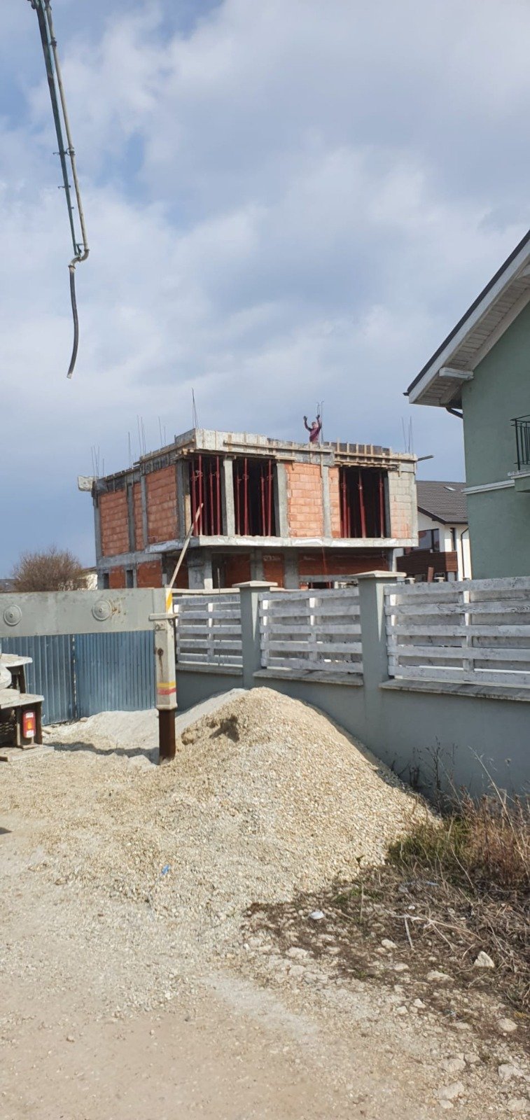 Construction site with a partially built brick house, construction materials and gravel in the foreground, and a person on the roof waving.