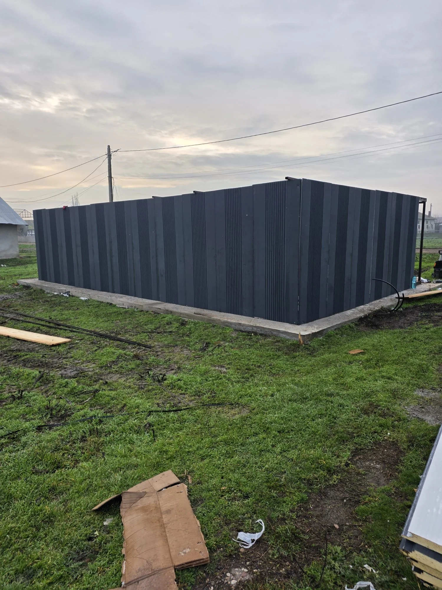 A rectangular building with black metal siding on a concrete slab in a grassy area, with an overcast sky in the background.