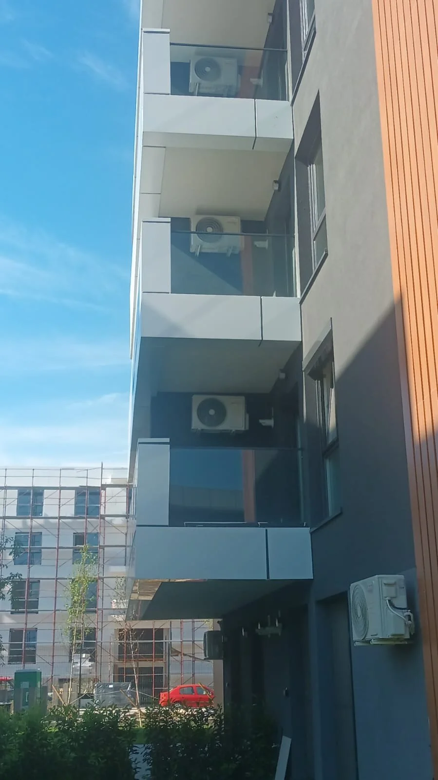 Three modern apartment balconies with glass railings and air conditioning units, seen against a clear blue sky, with nearby buildable area and a red car parked outside.