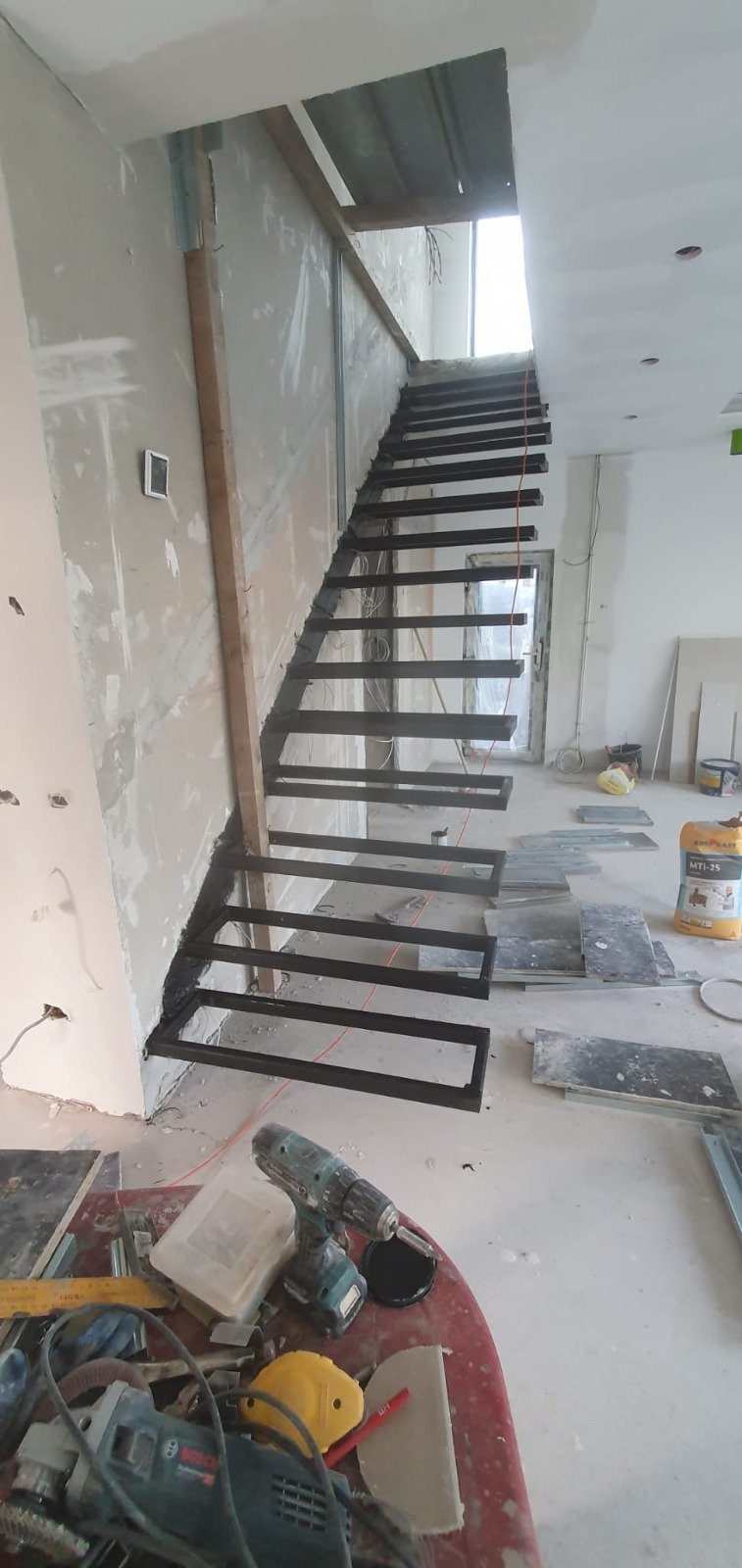 Construction site showing a staircase under construction with black metal steps and wooden supports, surrounded by tools and construction materials.