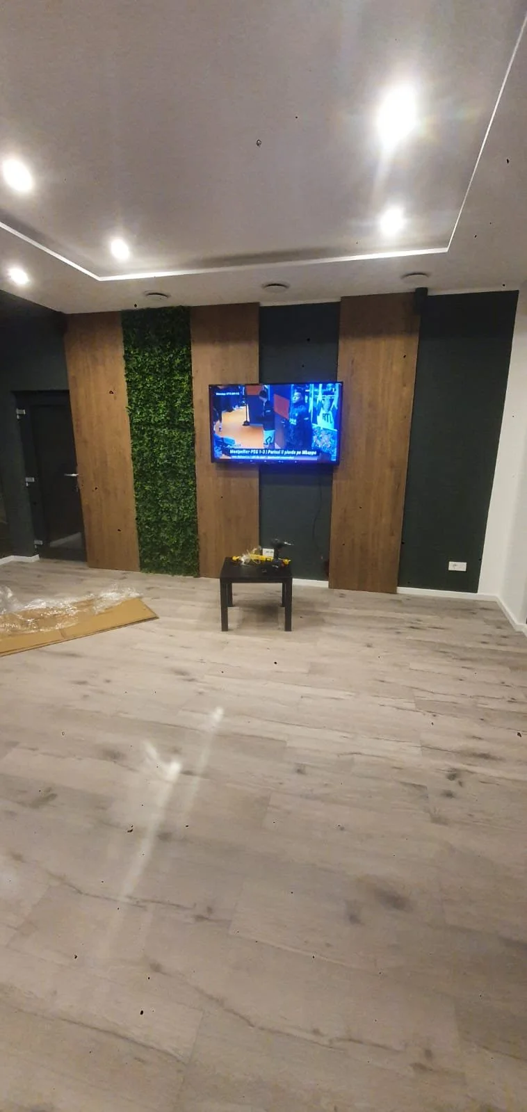 Empty room with a mounted TV on a wooden and dark wall, small black table with tools, and a partially unpacked cardboard box on the floor.
